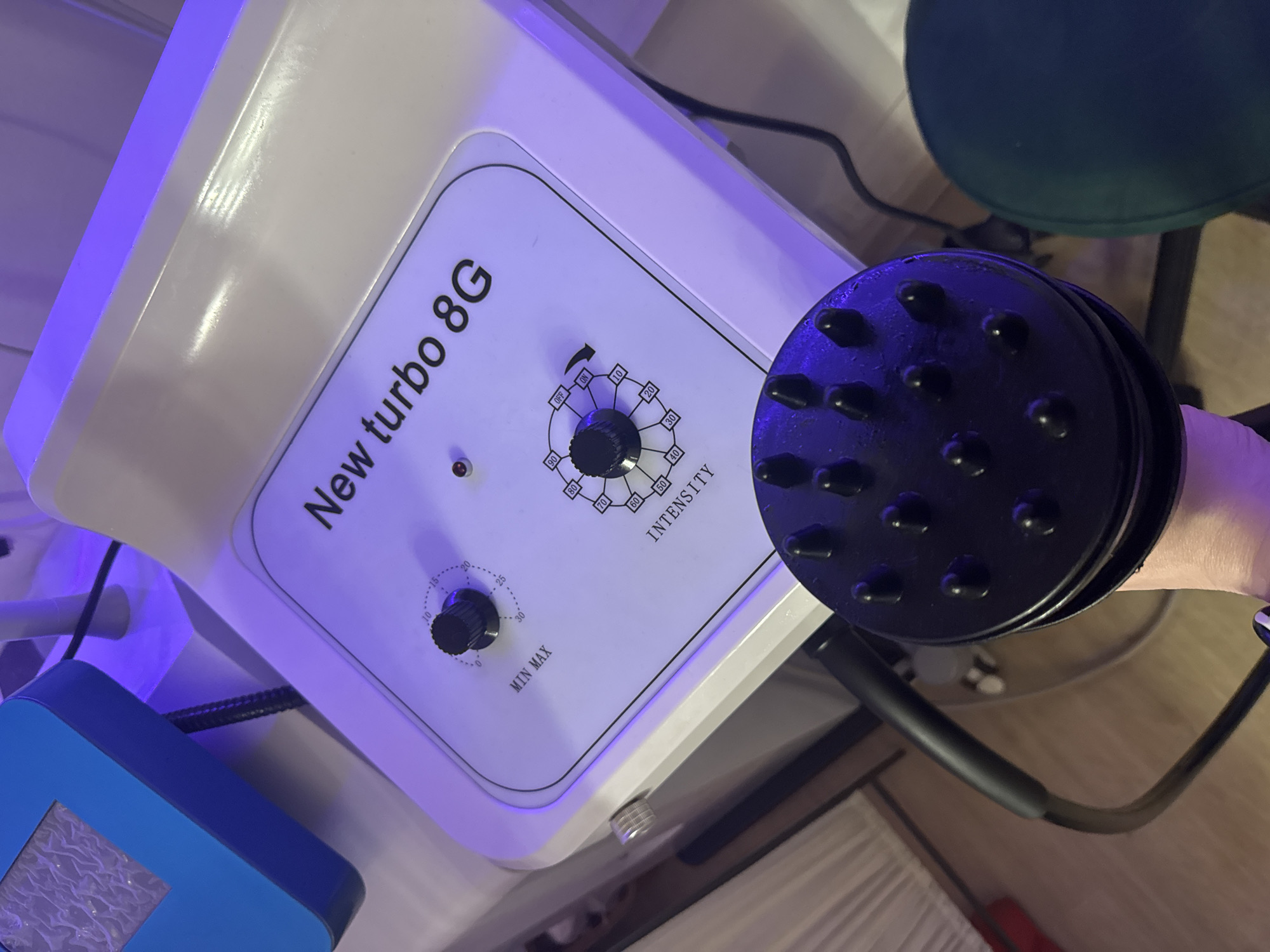 The image shows a white electronic device with "New turbo 8G" printed on the front panel. The device features two black control knobs and an intensity scale with numerical markings. A black handheld applicator with multiple rounded protrusions is connected to the device via a black cable. A blue and white rectangular object is visible on the left side of the image. The background includes a green chair and a light-colored floor. The setting appears to be indoors. There's no information to suggest the location is Saint Petersburg, Russia.