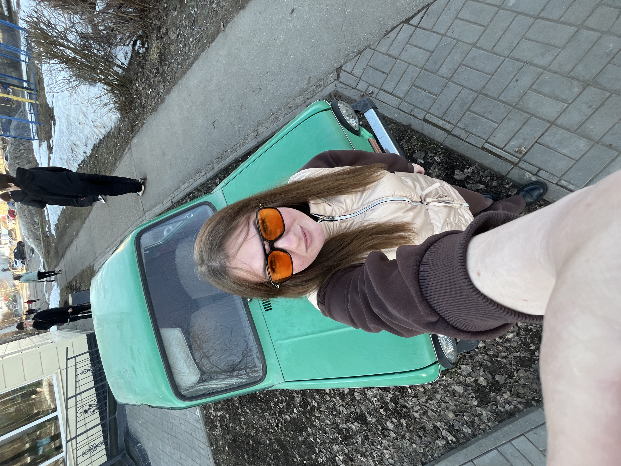 A young woman with long brown hair and orange-tinted sunglasses poses for a selfie outdoors, leaning against the hood of a faded green car. She wears a tan puffer vest over a brown sweatshirt. The car is parked on a paved surface beside a curb. Fallen leaves litter the ground around the car. In the background, a row of buildings, including a light-colored house with a black wrought-iron fence, is visible. Several people walk along the sidewalk in the distance. In the far background, a playground and bare trees can be seen. The location is Chaikovsky, Russia.