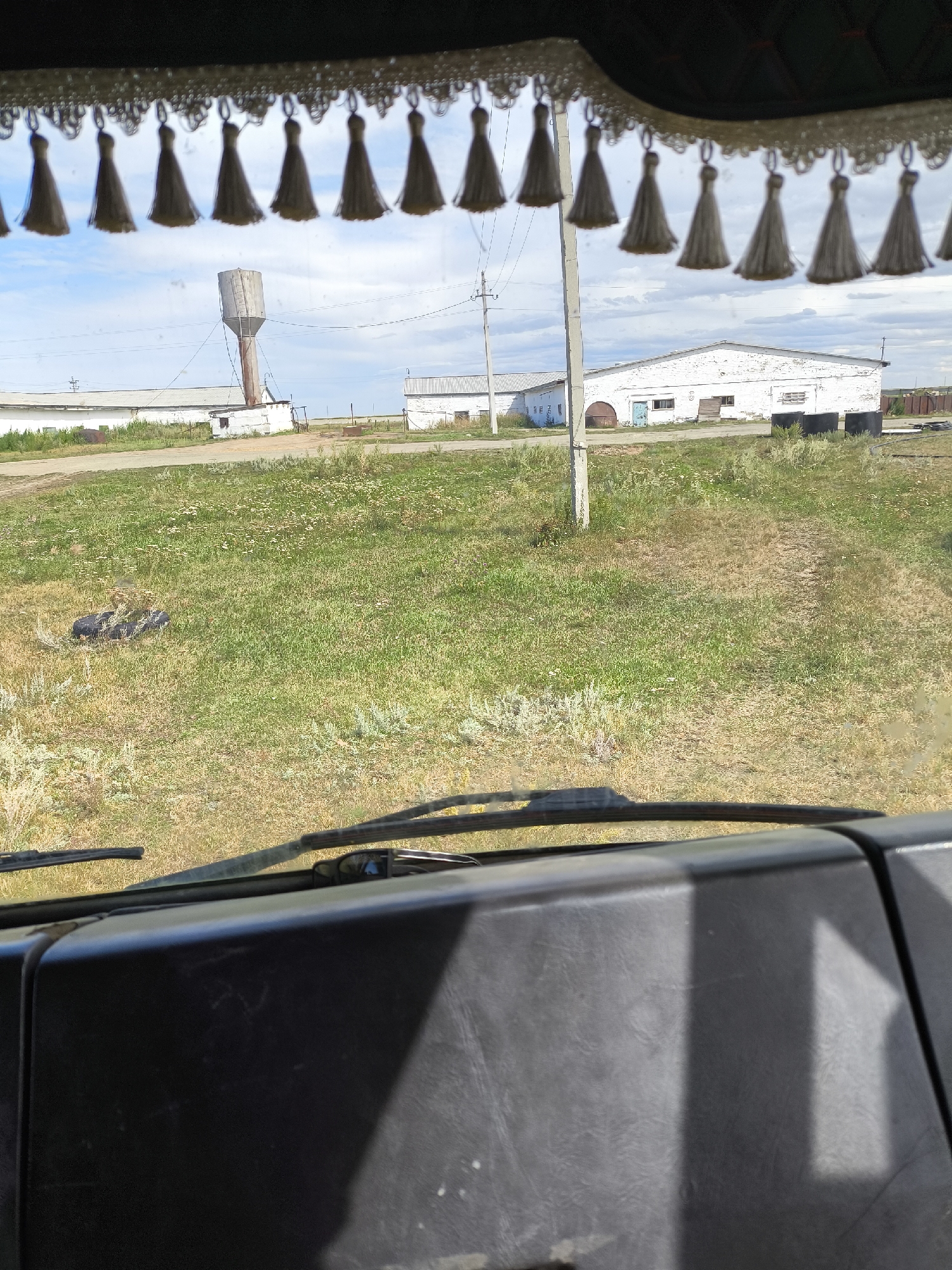 A low-angle, forward-facing shot from inside a vehicle shows a rural industrial scene under a cloudy sky. In the foreground, the dark, textured dashboard and windshield wipers are visible. Beyond the windshield, a wide expanse of dry, grassy fields dotted with small white wildflowers stretches towards a cluster of white industrial buildings and a tall, weathered water tower. A utility pole stands near the buildings. The scene is framed by a decorative fringe of grey tassels hanging from the top of the windshield. The environment appears to be an agricultural or industrial area in the city of Pripyat, Kazakhstan.