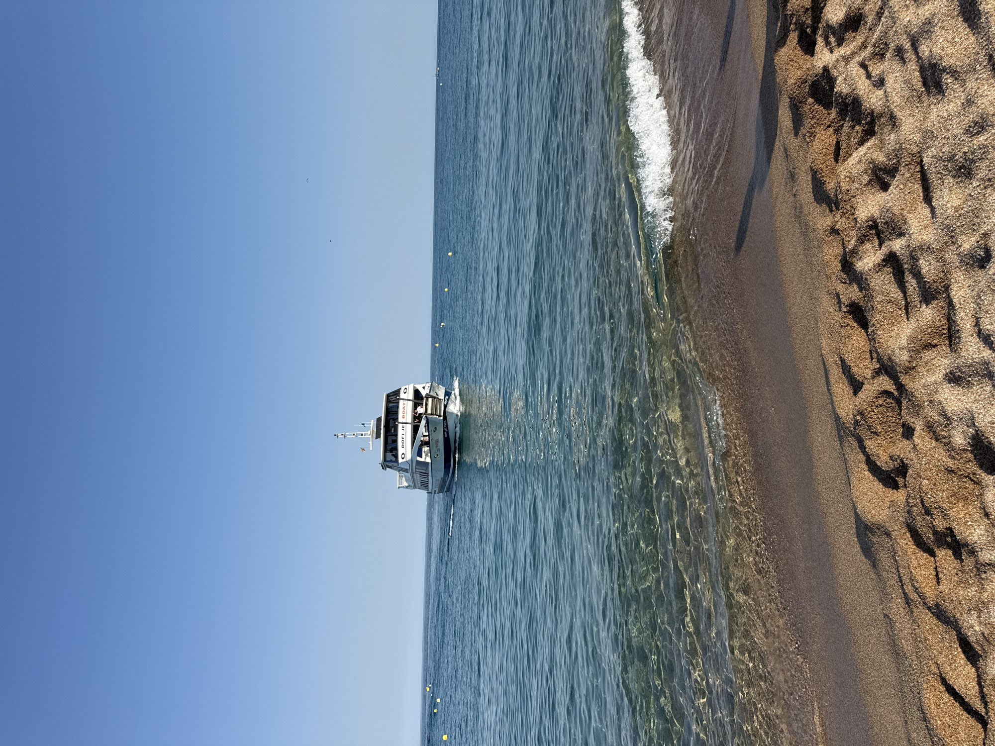 The image shows a boat sailing in the clear blue waters of the Mediterranean Sea off the coast of Blanes, Spain. The sandy beach is visible in the foreground, with gentle waves lapping at the shore. The sky is a clear, bright blue, indicating a sunny day. The boat, labeled "DOFI JET BOAT", appears to be a tourist vessel, possibly offering excursions. The water is calm and reflects the sunlight, creating a shimmering effect. There are a few yellow buoys floating in the sea, marking a designated area. The scene evokes a sense of peace and relaxation, characteristic of a summer day at the beach. The photograph captures a moment of leisure and enjoyment in a beautiful coastal setting. The time of day appears to be late morning or early afternoon, given the bright sunlight and the angle of the shadows.