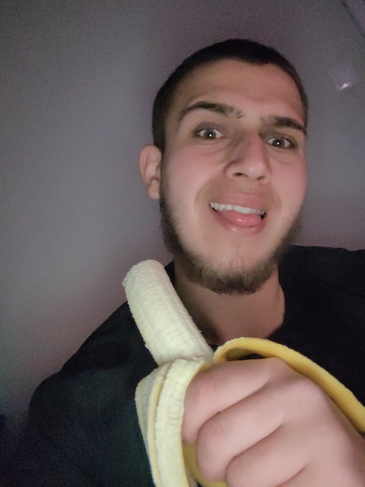 A man is holding a peeled banana in his left hand. The man has short brown hair and a beard. He is wearing a black shirt. The man is sticking out his tongue. The location is not discernible from the image. There is no indication that the picture was taken in Sofia, Bulgaria.