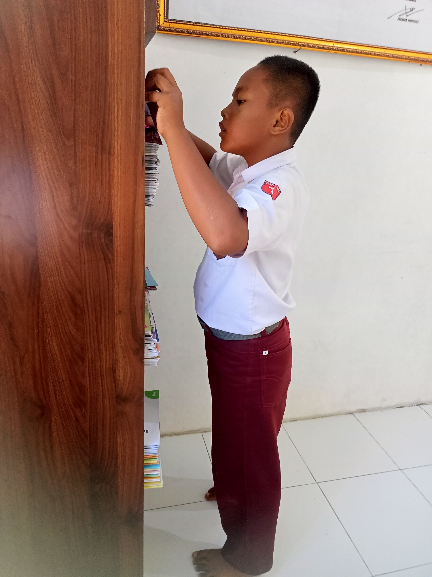 In Kagungan Ratu, Indonesia, a young boy stands next to a wooden shelf, reaching for a stack of books. He is wearing a white short-sleeved shirt with a red emblem on the left chest and dark red pants. The shelf is made of polished wood and appears to be part of a larger piece of furniture. The wall behind him is a plain white. The floor is tiled with white squares. The boy's feet are bare. The lighting suggests an indoor environment, possibly a school or library.