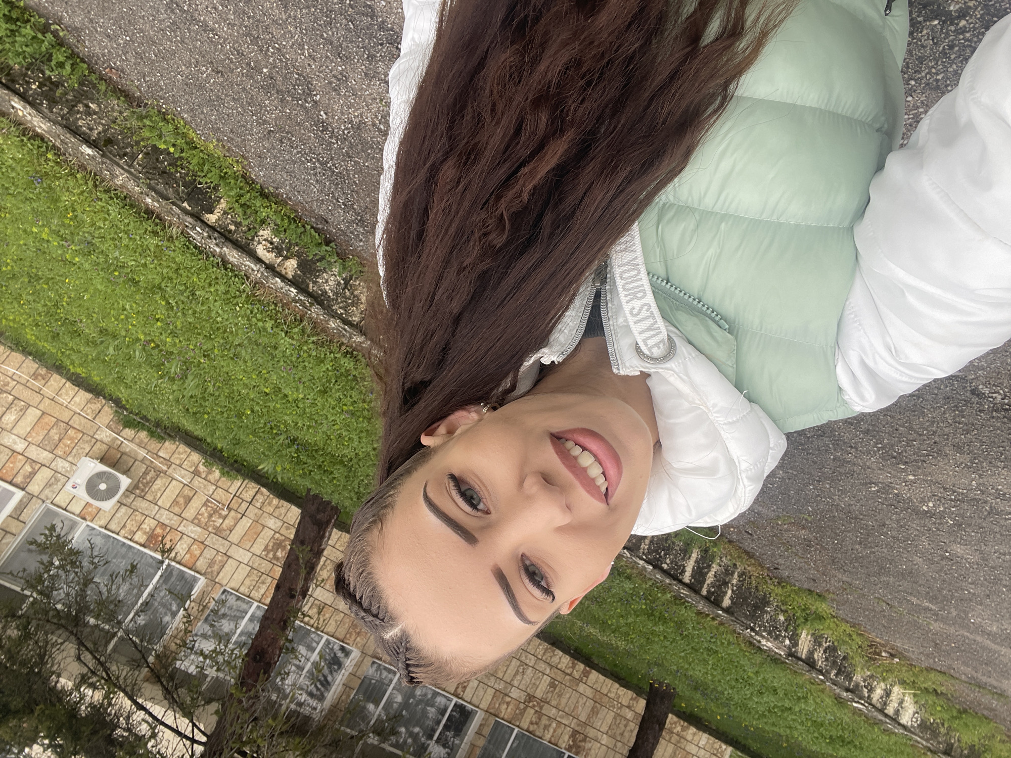 The image shows a young woman with long, dark brown hair wearing a white and light green puffer jacket. She is smiling and looking directly at the camera. The background includes a building with brick walls and windows, an air conditioning unit, and some trees. There is also a sidewalk and a grassy area visible. The location is suggested to be Veliko Tarnovo, Bulgaria.