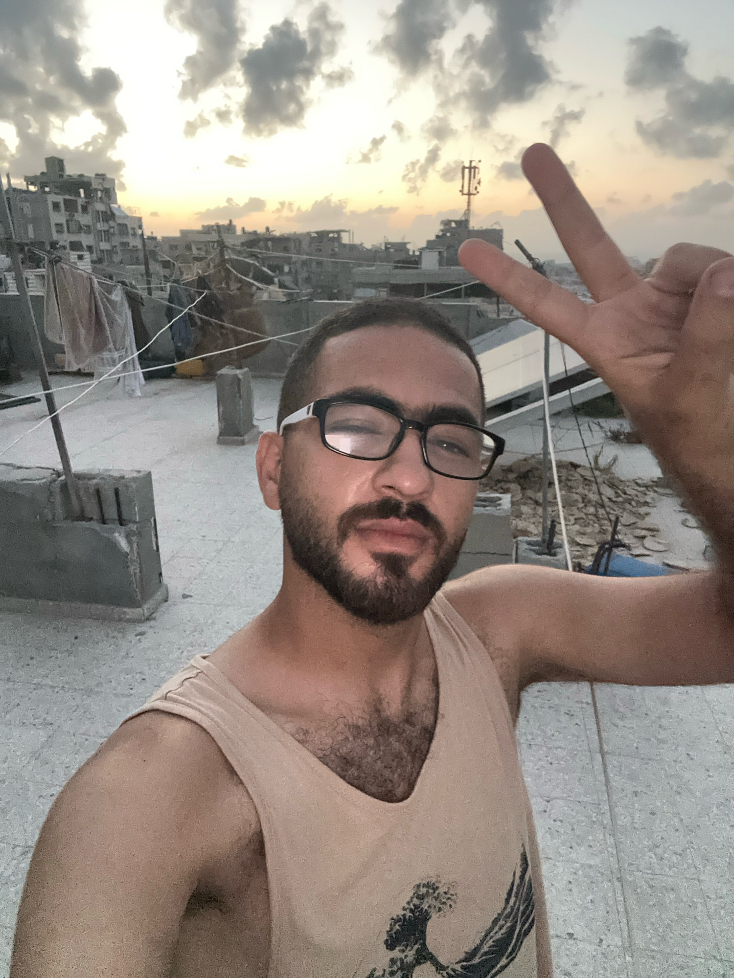 An outdoor, selfie-style medium shot captures a young adult male from the chest up, standing on a rooftop with an urban Gaza City skyline visible in the background under a cloudy sky. The man is wearing glasses, a light-colored tank top, and has a beard. He is making a peace sign with his right hand. The rooftop is made of concrete and appears to have some building materials and laundry hanging on lines. The cityscape consists of multi-story buildings, some appearing worn, and a telecommunications tower. The lighting suggests it is either sunrise or sunset.