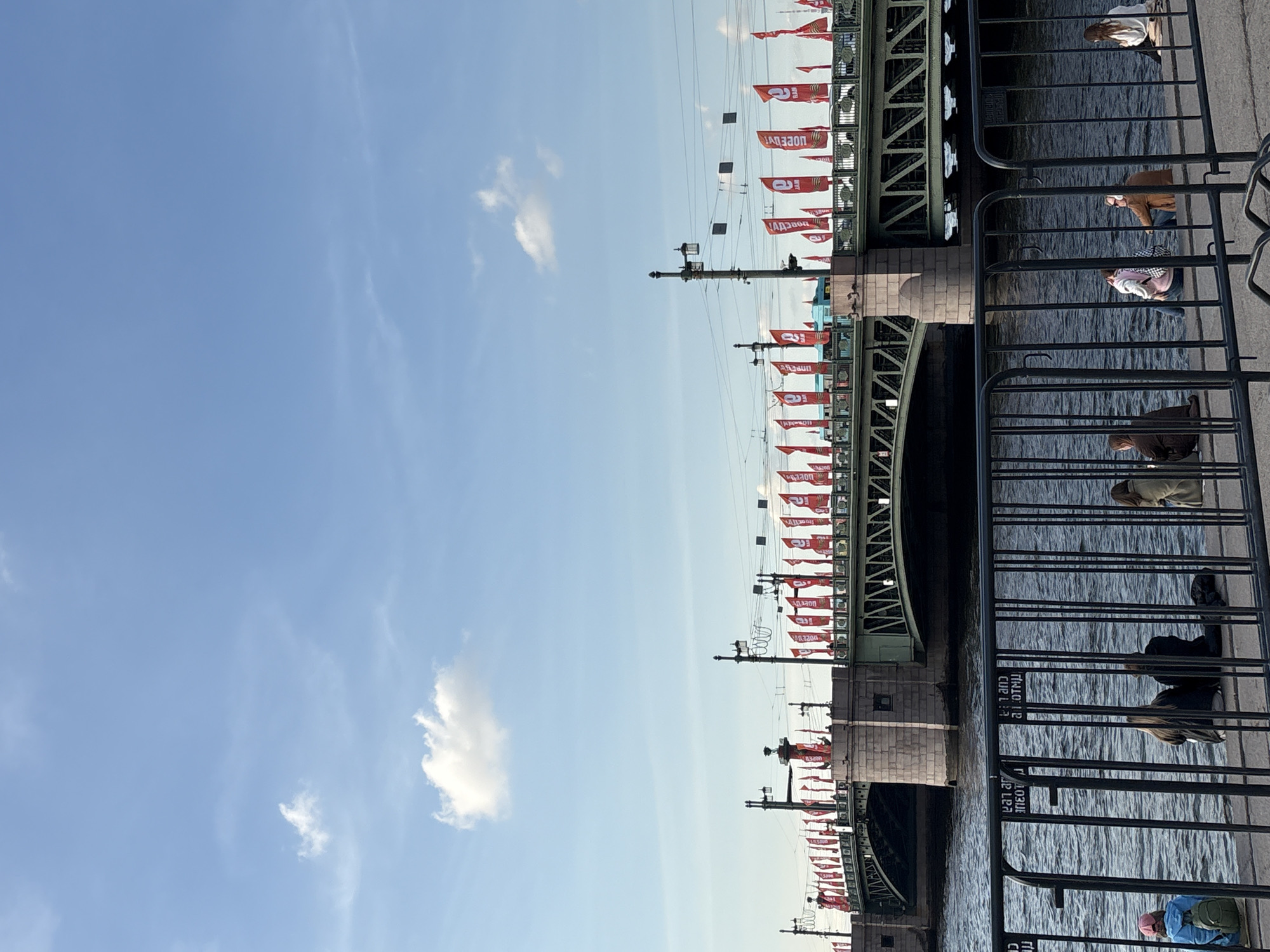The image depicts a bridge in Saint Petersburg, Russia, adorned with numerous red banners displaying white Cyrillic text. The bridge's architecture features a dark green metal framework and stone support structures. Pedestrian barriers line the edge of the bridge, separating the walkway from the water below. Several people are visible on the bridge, leaning against the barriers and looking out at the water. The sky is clear and blue, with a few scattered white clouds. The water beneath the bridge is calm and reflects the sunlight. The scene appears to be captured during the daytime.