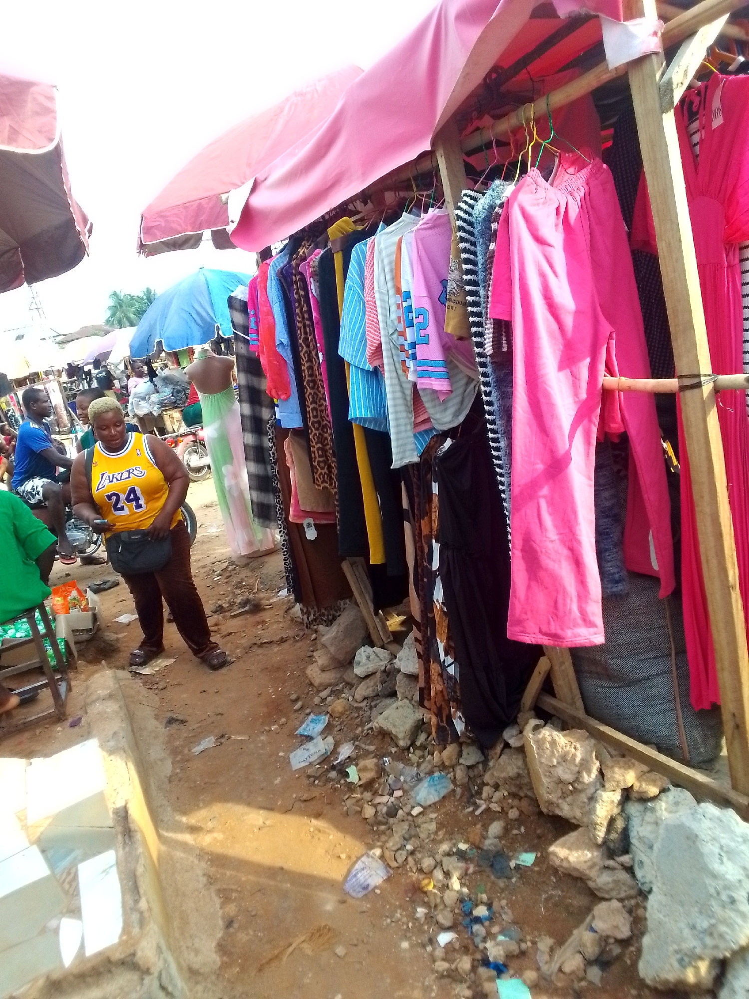 The image depicts a street scene in Orlu, Nigeria. A woman wearing a yellow Lakers jersey with the number 24 and brown pants stands in front of a rack of clothing for sale. The clothing rack is constructed of wood and covered with a pink canopy. Various garments in shades of pink, blue, black, and patterned designs hang from the rack. Debris, including rocks and discarded packaging, litters the ground beneath and around the clothing rack. In the background, other people and market stalls are visible, covered by umbrellas or tarpaulins. A man is riding a motorcycle.