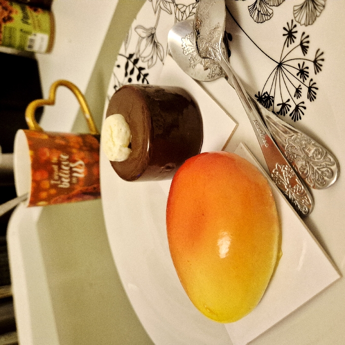 A dessert plate features a cylindrical chocolate dessert topped with white cream next to a vibrant orange and yellow oval dessert. Two ornate silver spoons rest diagonally across the plate, which has a floral pattern around the edge. A mug with a heart-shaped handle and the phrase "I need to believe in us" is visible. Part of a jar is also seen in the background. The setting suggests a casual, perhaps domestic, environment.