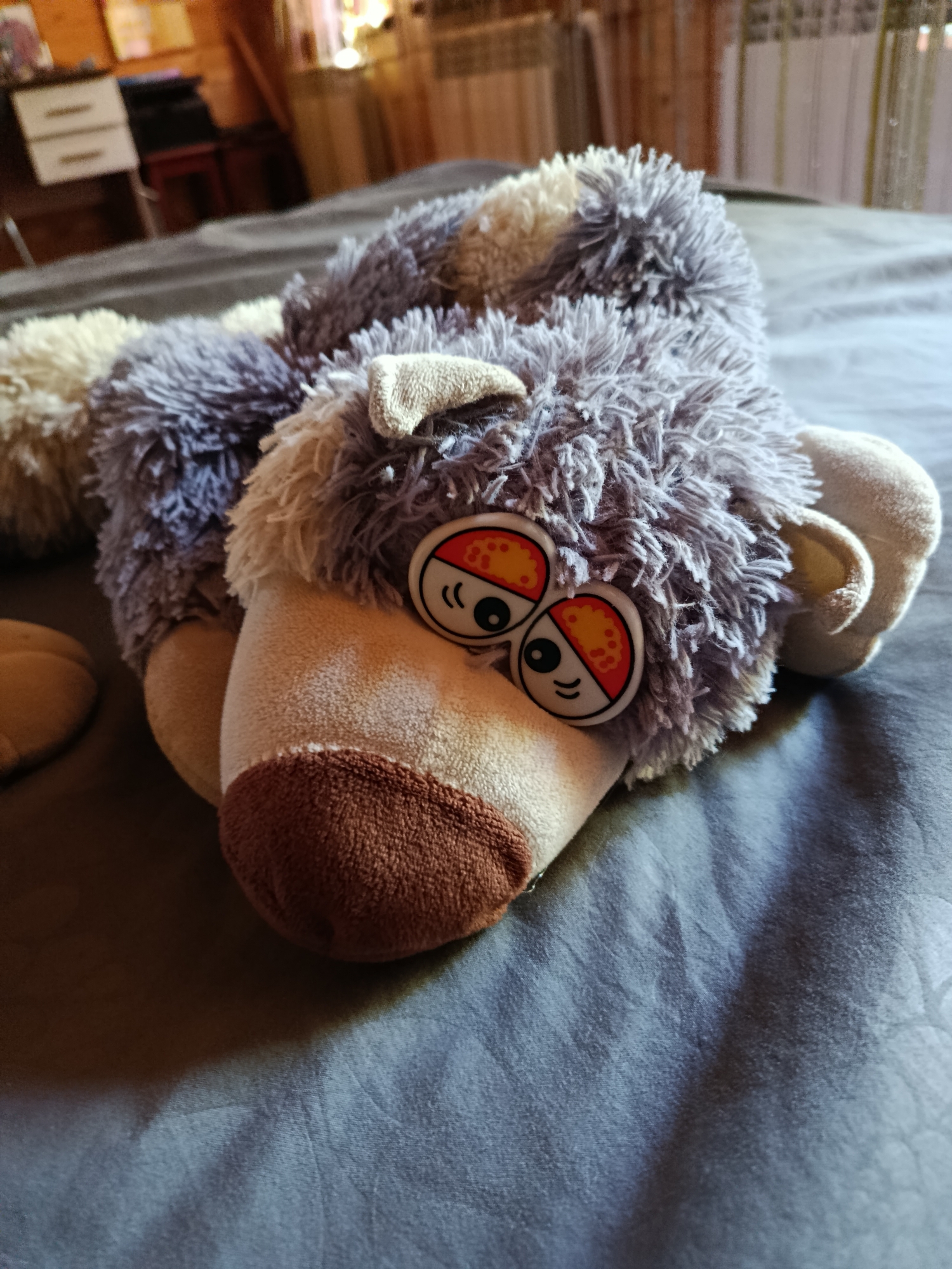 A multi-textured plush toy, predominantly grey with beige accents and shaggy fur, lies horizontally on a dark grey, subtly wrinkled fabric surface. The toy features two large, circular, cartoon-style eyes, each divided into an upper orange half with white dots and a lower white half with a black pupil and two black curved lines. A prominent brown snout is centered below the eyes. Small, beige, floppy ears are visible on either side of the head, and a beige paw is partially visible on the left side. The background is out of focus, showing a domestic interior with light brown wooden paneling, a white two-drawer cabinet on the left, and a dark furniture item. The scene is illuminated by soft, diffused light. This image was captured in the city of Vereya, Russia.