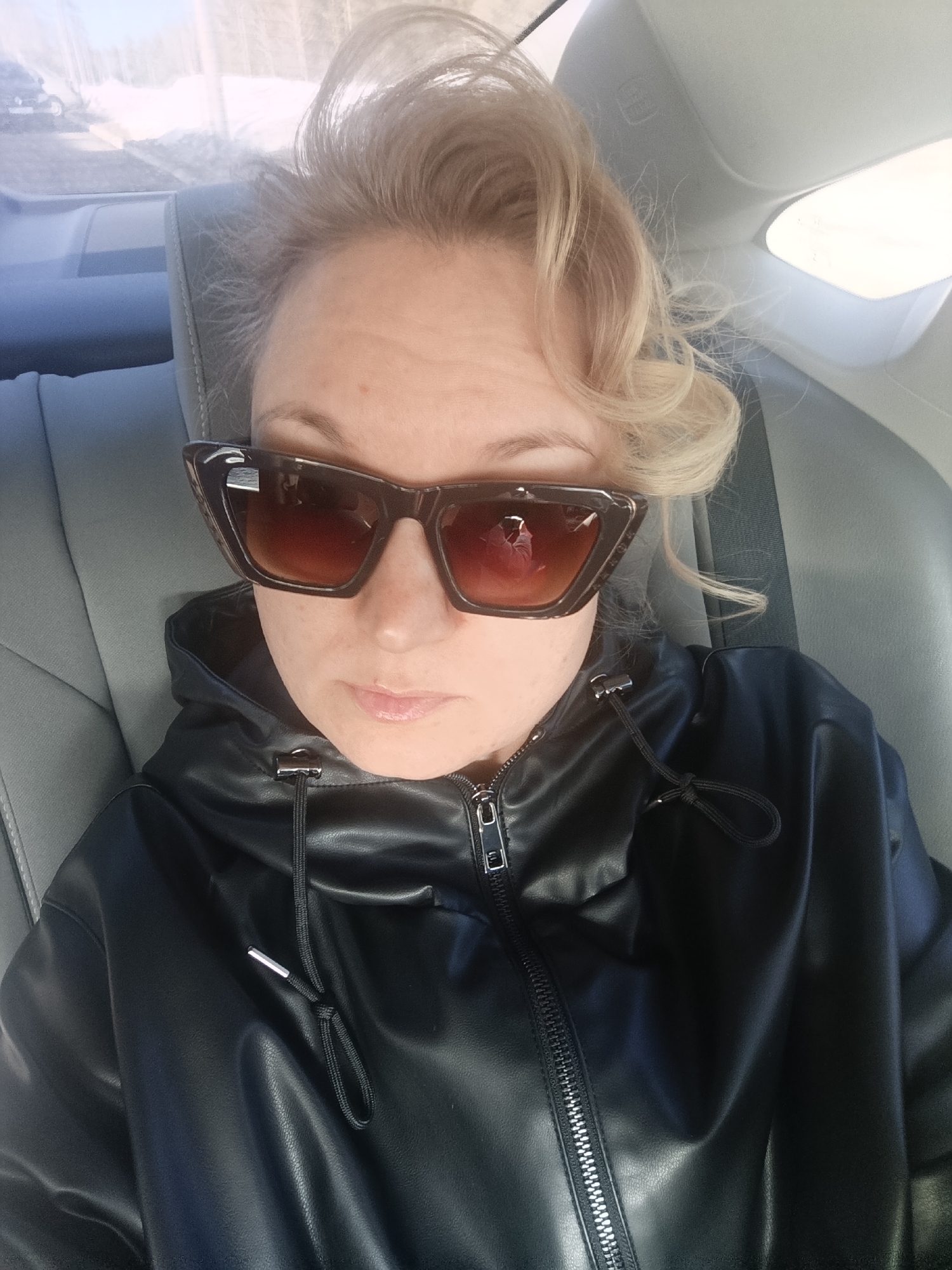The image depicts an individual seated in the gray interior of a vehicle, located in the city of Марковский, Russia. The subject, an adult with blonde hair, is wearing dark, cat-eye style sunglasses with amber-tinted lenses and a black faux leather hooded jacket. The jacket features a visible front zipper and a drawstring with silver-colored tips. A reflection of a person holding what appears to be a camera or phone is observable in the left sunglass lens. The vehicle's gray upholstered seat and headrest are visible behind the subject. Through a window, a blurred exterior environment consistent with a winter landscape, including snow and bare vegetation, is discernible under daylight conditions.