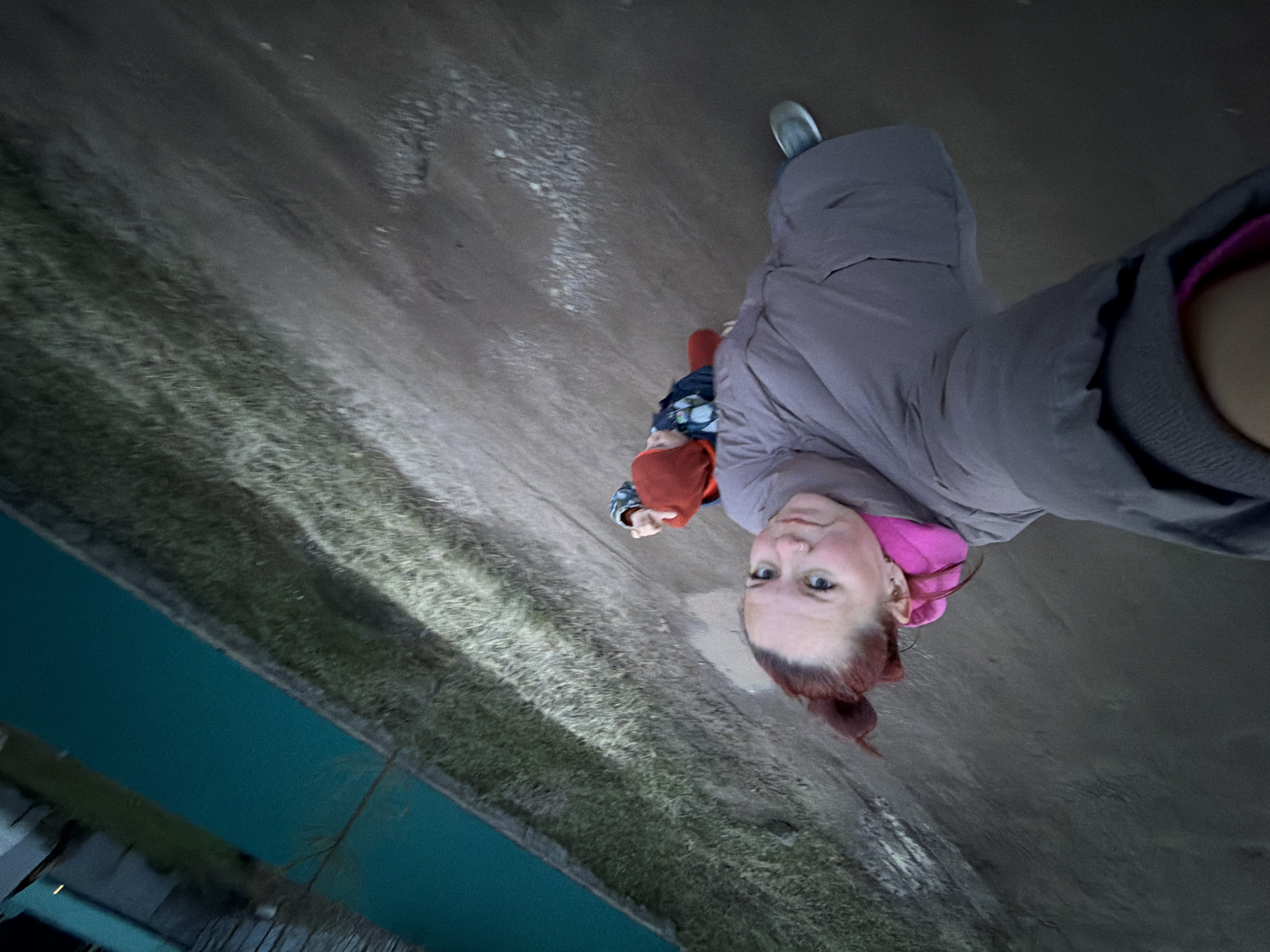 A low-angle, rotated selfie captures a woman and a child outdoors in what appears to be Chaikovsky, Russia. The woman, positioned in the foreground on the right, looks directly at the camera with a neutral to slightly curious expression. She has reddish-brown hair tied back and is wearing a gray puffer jacket with a bright pink collar or inner layer visible. A silver nose ring is visible.
Behind her, and slightly to the left, a small child is partially visible, wearing a red hat and a dark blue or black patterned jacket, walking away from the camera. The ground beneath them is a dark, uneven surface, possibly a dirt path or muddy area, with patches that look wet or icy. To the left, a strip of green, textured grass runs alongside a calm, distinctly turquoise-blue body of water, possibly a canal or river. The lighting is muted and ambient, suggesting an overcast day or twilight. There is no visible text.
