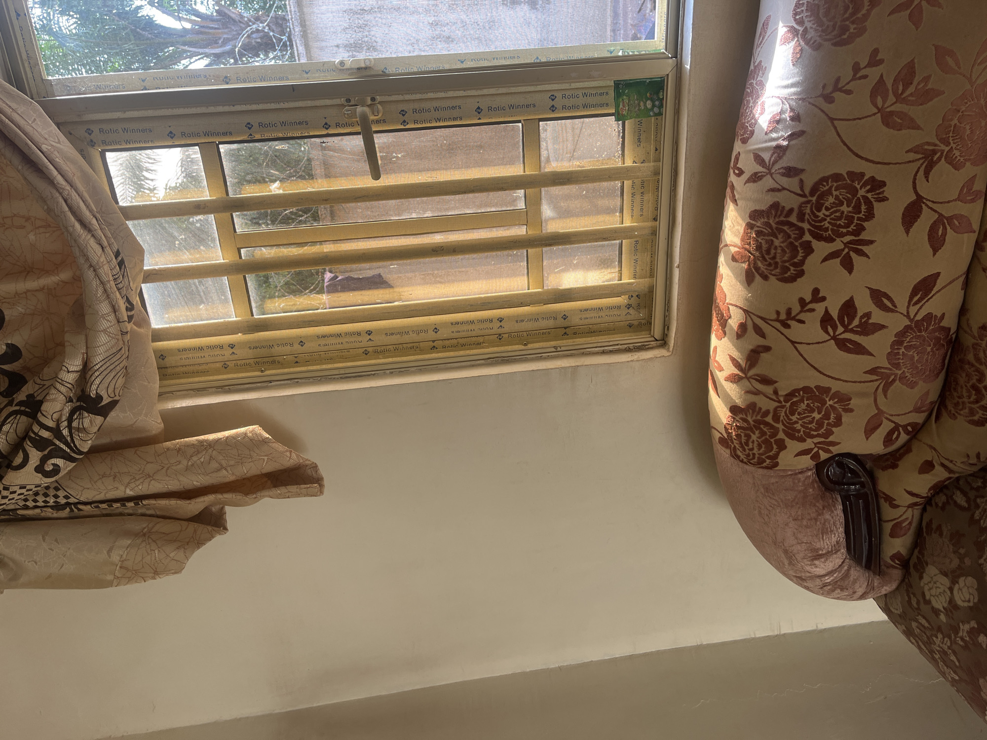 The image shows an interior scene, likely a room, with a window on the left and a sofa on the right. The window has a metal grille and is covered by sheer curtains, one of which is pulled to the side, revealing a slightly distorted view of trees and sunlight outside. The window frame is adorned with repeating text that reads "Rotic Winners". The sofa is upholstered in a floral fabric with a dominant pattern of dark red roses and leaves on a light beige background. Part of the sofa's armrest is visible, featuring a dark, ornate wooden detail. Sunlight streams in from the window, casting shadows and highlighting the textures of the curtains and sofa. The overall impression is one of a domestic setting, possibly in the daytime, given the presence of sunlight. The repeating "Rotic Winners" text suggests branding or decorative elements on the window. The image's orientation is slightly tilted, giving a dynamic perspective to the scene.