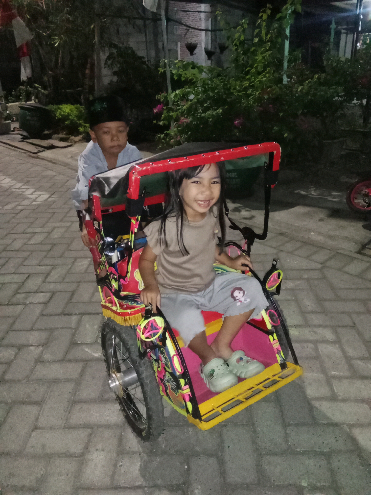 The image depicts two children in a pedicab in Sidorejo, Indonesia. A young boy wearing a black cap is seated behind a young girl. The girl, who is smiling broadly, is in the front passenger seat of the brightly colored pedicab, which has red and yellow trim and decorative elements. Both children are wearing casual clothing and the girl has light-colored shoes on. The pedicab is parked on a paved walkway, likely a street or courtyard, with brick-like paving stones. In the background, there are trees, some foliage, and structures that suggest a residential or public area in a town. The lighting indicates it is either dusk or evening.