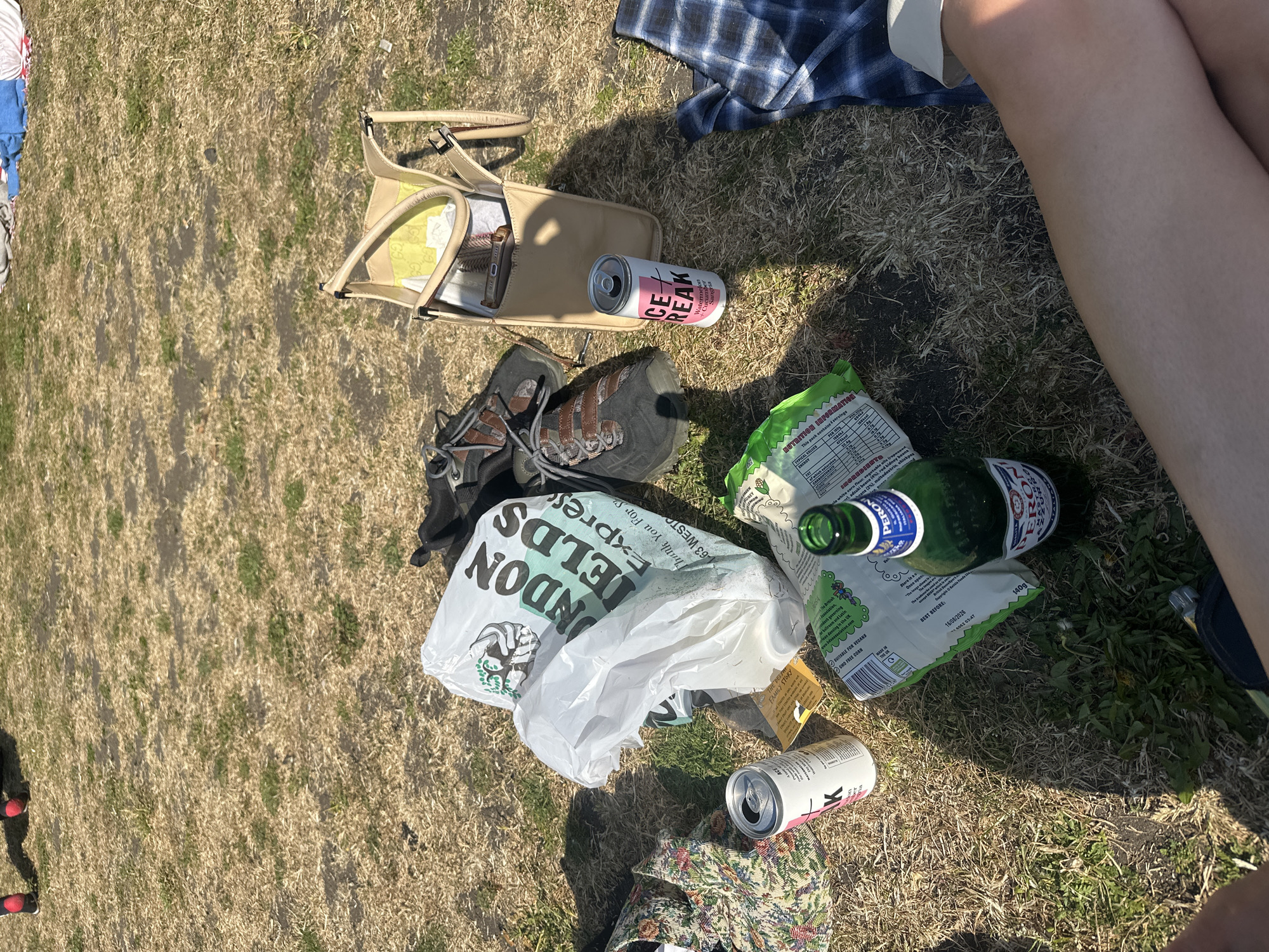 The image shows a grassy area, possibly a park or field, with various discarded items. There's a handbag lying open, next to a hiking boot and a "LONDON FIELDS Express" plastic bag. The scene includes an empty bottle of Peroni beer, a crumpled bag of crisps, and two cans of "ICE BREAK" watermelon and cucumber-flavored sangria. A glimpse of a person's legs is visible in the upper right corner. Given the text on the bag, the location is likely somewhere near London Fields in the Hackney area of London, United Kingdom.