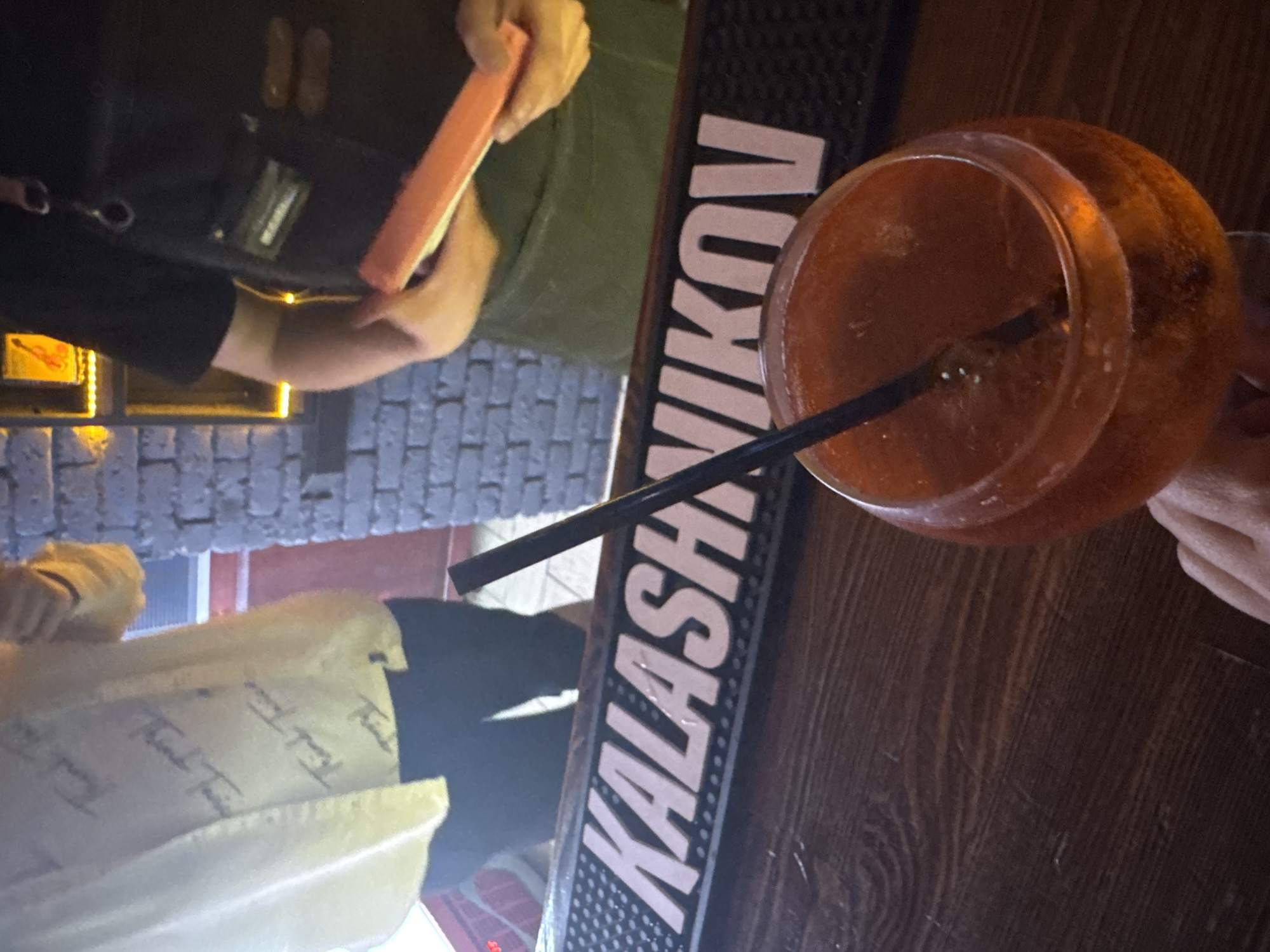 The image shows a dimly lit bar scene. A person wearing a light-colored jacket with printed text and dark pants is standing on the left side of the frame. Adjacent to this person is a black surface with white lettering that spells "KALASHNIKOV". On the right, there's a close-up of an orange-colored cocktail in a round glass, with a black straw extending into the drink. A person is holding the cocktail glass. Another person in black clothing is seen in the upper-left, holding a rectangular, possibly pink, object. The background features a brick wall with dim lighting. The location is the city of Chaikovsky, Russia.