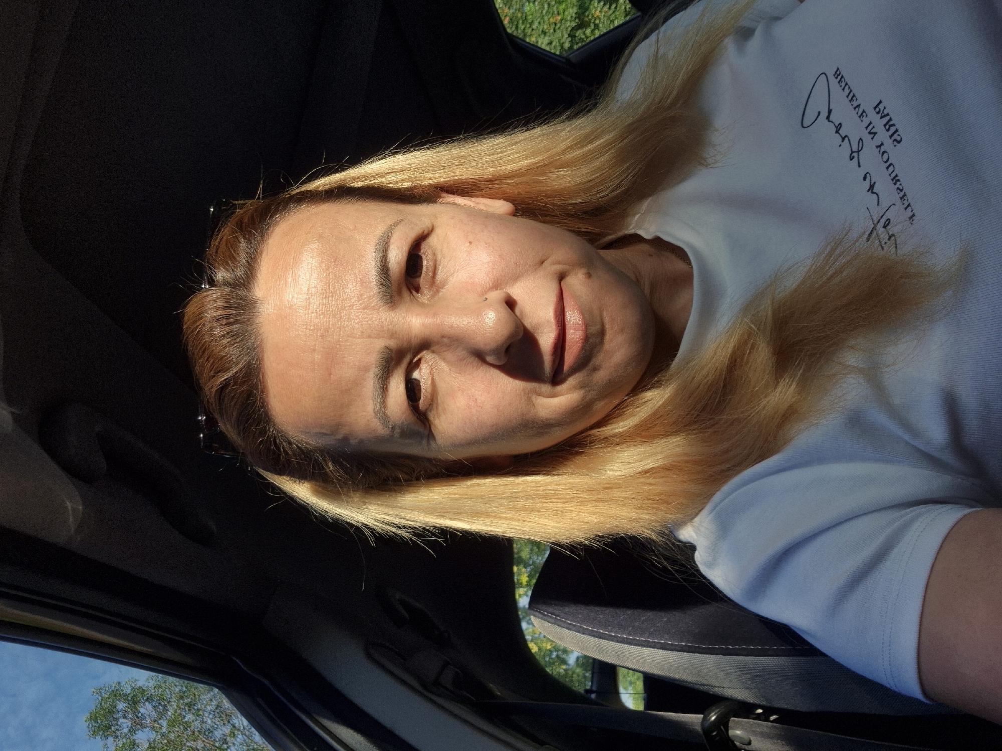 This is a vertical, close-up, selfie-style photograph taken from inside a vehicle. The primary subject is a woman with shoulder-length, blonde hair. She is looking towards the camera with a slight smile and her head is tilted. Sunlight is casting a strong shadow on the left side of her face. She is wearing a light-colored t-shirt with some text printed on it. The text appears to be in a stylized font and is in a dark color. The background is partially visible, showing the interior of a car with a dark dashboard and a glimpse of green foliage and blue sky through a window. The lighting suggests it is daytime, likely late afternoon due to the harsh shadows. The overall mood appears casual and relaxed.