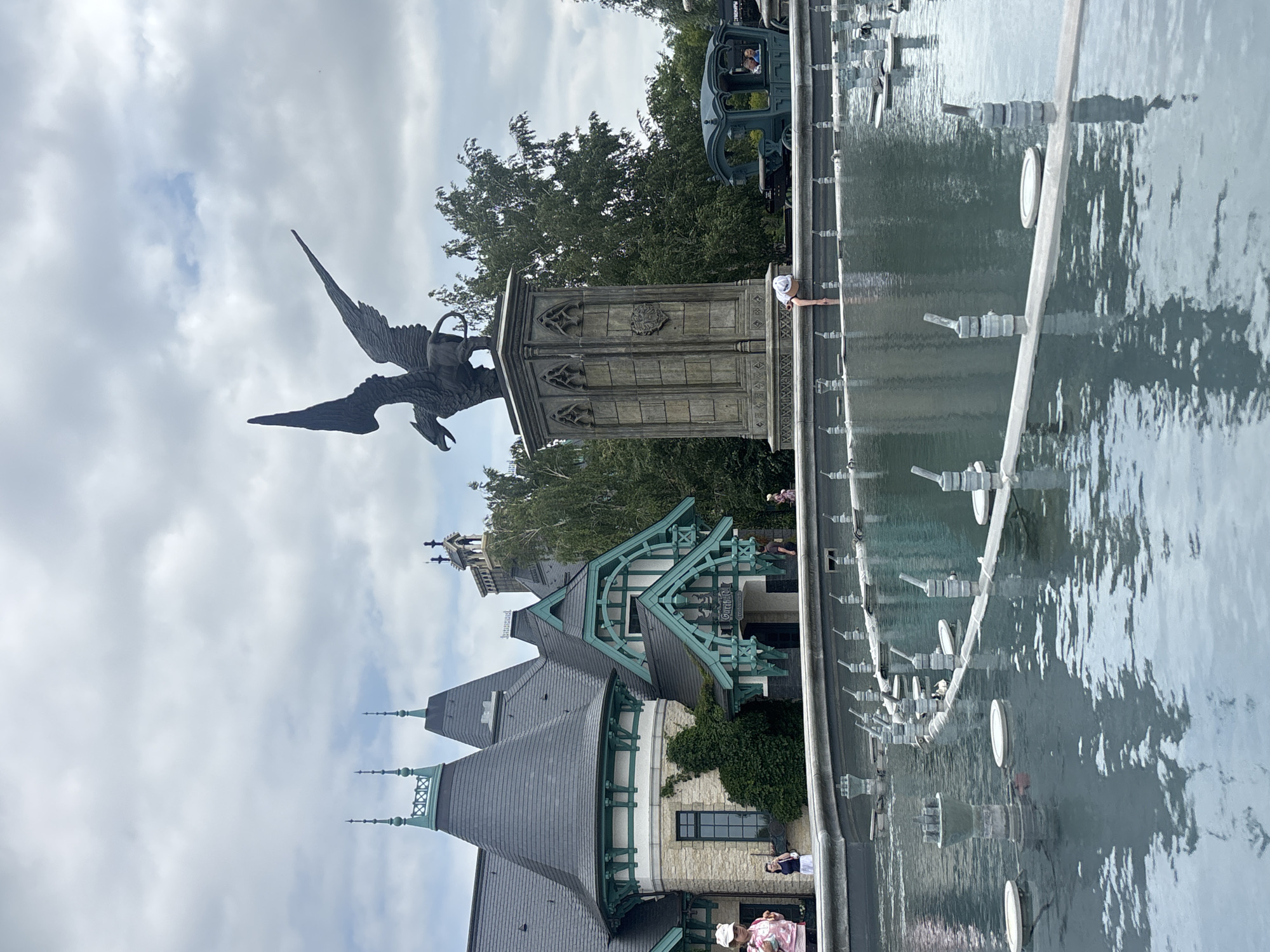 The image captures a view of the Fountain of Khryashchevka, located in Khryashchevka, Russia. In the foreground, a large, ornate fountain with many spouts is partially filled with water, reflecting the cloudy sky and surrounding structures. A person is seen reaching into the water from the edge of the fountain. In the middle ground, a prominent statue of a griffin stands atop a stone pedestal. To the right of the statue, a horse-drawn carriage with passengers is visible. Behind the fountain and statue, a grand building with distinctive architecture, featuring pointed roofs and green trim, dominates the background. Trees surround the area. The sky is overcast with scattered clouds.