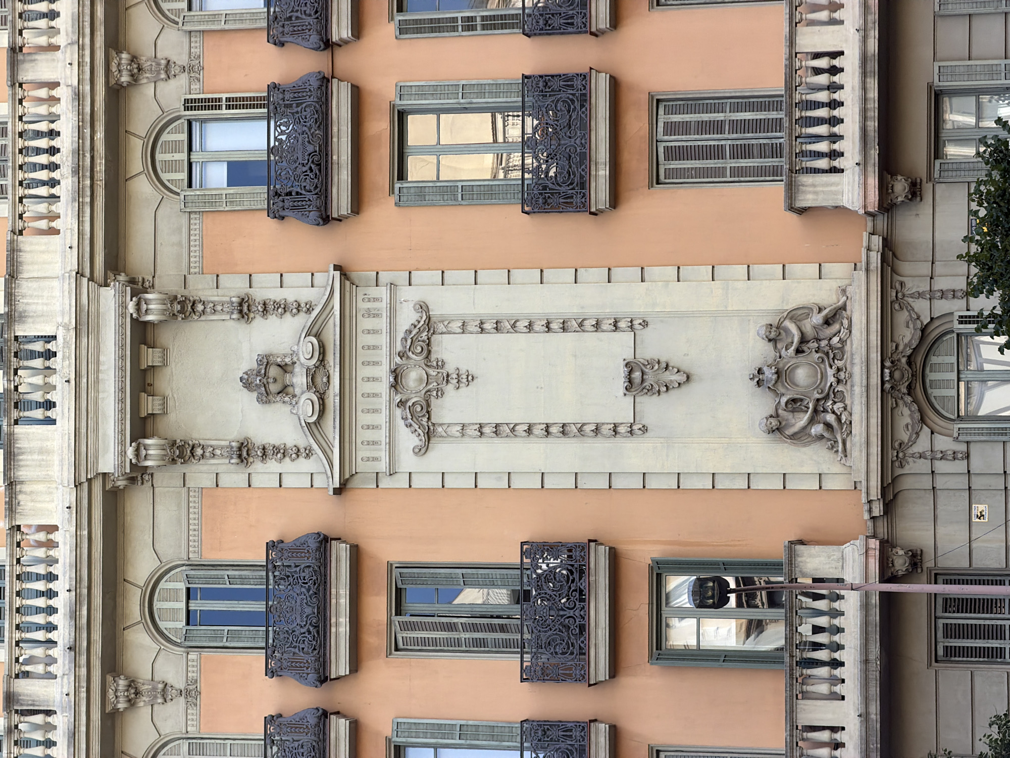 This is a close-up, head-on exterior architectural photograph of the facade of a building in Barcelona, Spain. The building features a warm, peach-colored stucco on the lower and mid-sections, contrasted with ornate, light-colored stone elements. There are multiple levels of windows, many with decorative wrought-iron balconies. The windows are framed by shutters, some of which are open, revealing reflections of the sky and surrounding architecture. The central portion of the facade showcases elaborate relief carvings, including a decorative urn and stylized floral motifs. In the lower part of the frame, there is a street lamp with a single light fixture. Green foliage is visible at the bottom left and bottom right of the image. The architectural style suggests a building from the late 19th or early 20th century, common in many European cities.