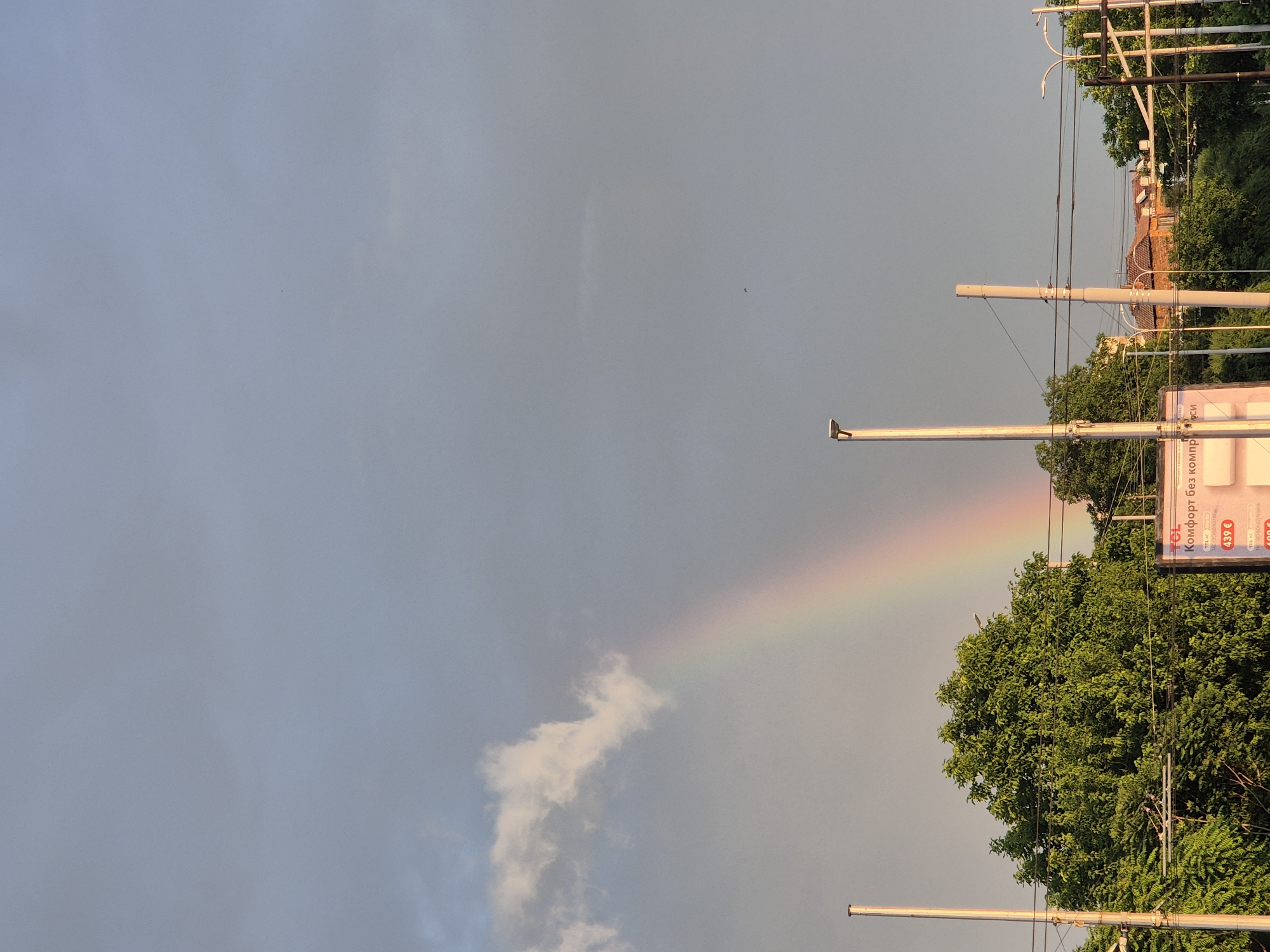 The image captures a vibrant rainbow arcing across a partly cloudy sky, indicating recent rainfall. The sky transitions from a soft grey-blue on the left to lighter tones on the right, with a distinct white cloud formation visible below the rainbow. A tiny, dark, unidentifiable speck floats in the upper left of the sky.
On the right side of the frame, urban or suburban elements are visible. Lush, dense green trees occupy the mid-ground, partially obscuring buildings with red-tiled roofs in the background. Several tall utility poles or streetlights with overhead wires stand prominently in front of the trees. A billboard is also partially visible on the far right.
The lighting suggests late afternoon or early evening, with a warm, golden glow illuminating the tops of the trees, building roofs, and the utility poles.
Visible text on the billboard, written in Cyrillic script, includes "Комфорт без ко..." (Comfort without co...), along with prices listed in Euros: "439 €", "499 €", and a partially visible "400 €". The letters "TOL.C" are also visible on the sign.