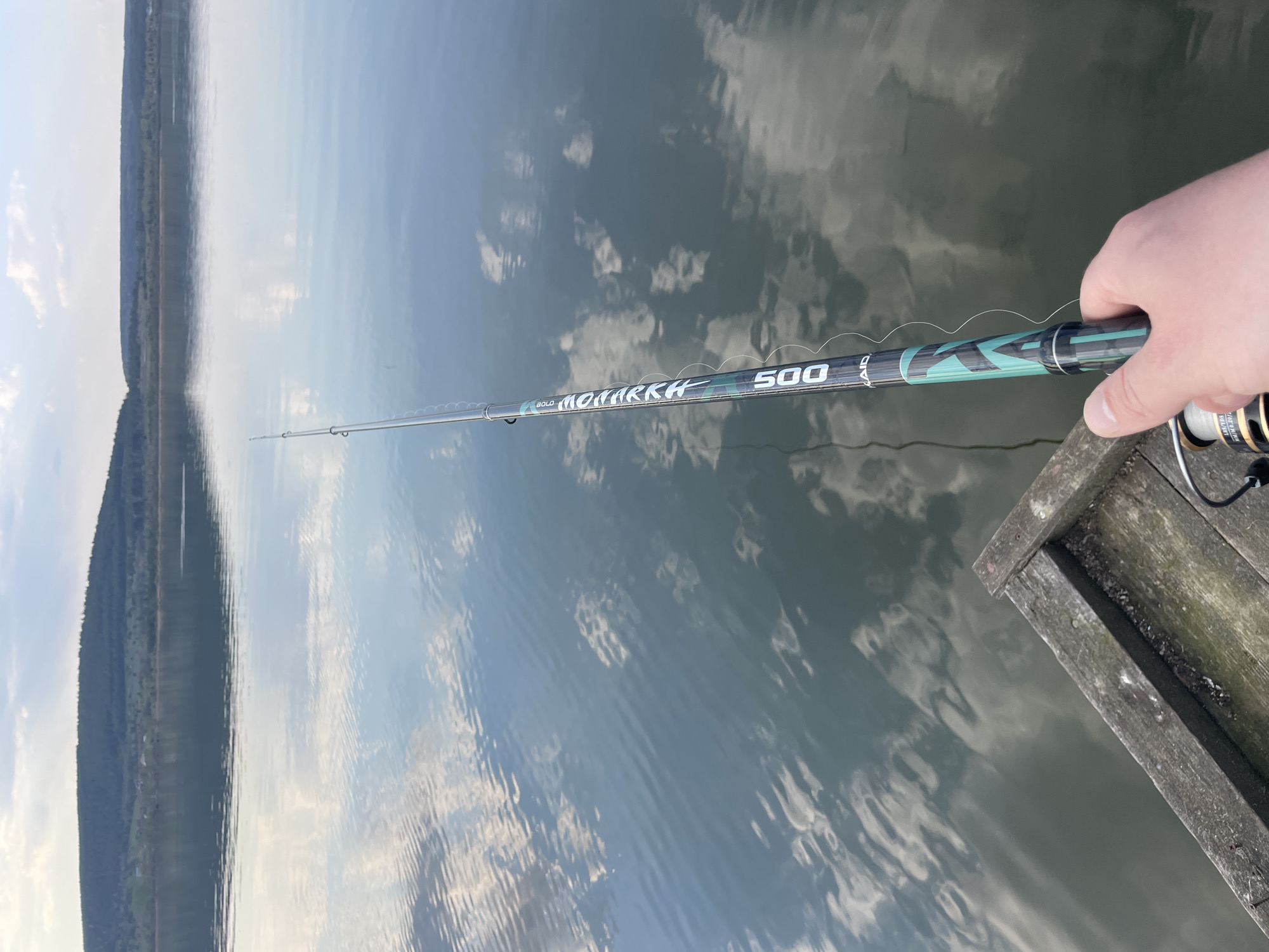 The image shows a fishing rod being held over a body of water, likely a lake or river, from a wooden dock or pier. The fishing rod is labeled "BOLO MONARKH 500". The background reveals a forested shoreline under a sky reflected in the water's surface. The location is identified as near Suksun, Russia.