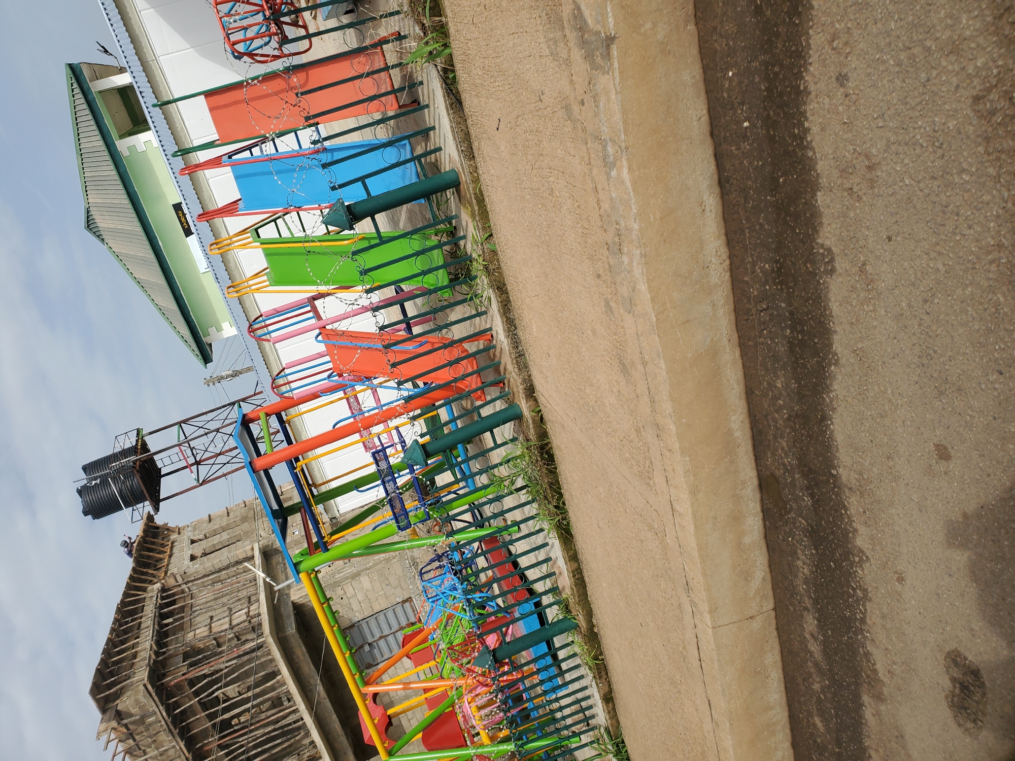 In Bukuru, Nigeria, a vibrant playground with various slides, swings, and merry-go-rounds in shades of red, blue, green, and yellow is visible. The playground is enclosed by a green metal fence adorned with barbed wire. Beyond the playground, a partially constructed building with wooden scaffolding stands against a cloudy sky, and a water tank on a metal platform is positioned nearby. To the right of the playground, a building with a green roof and white walls, possibly a school or community center, is also present. The ground in front of the playground is paved with concrete and asphalt.