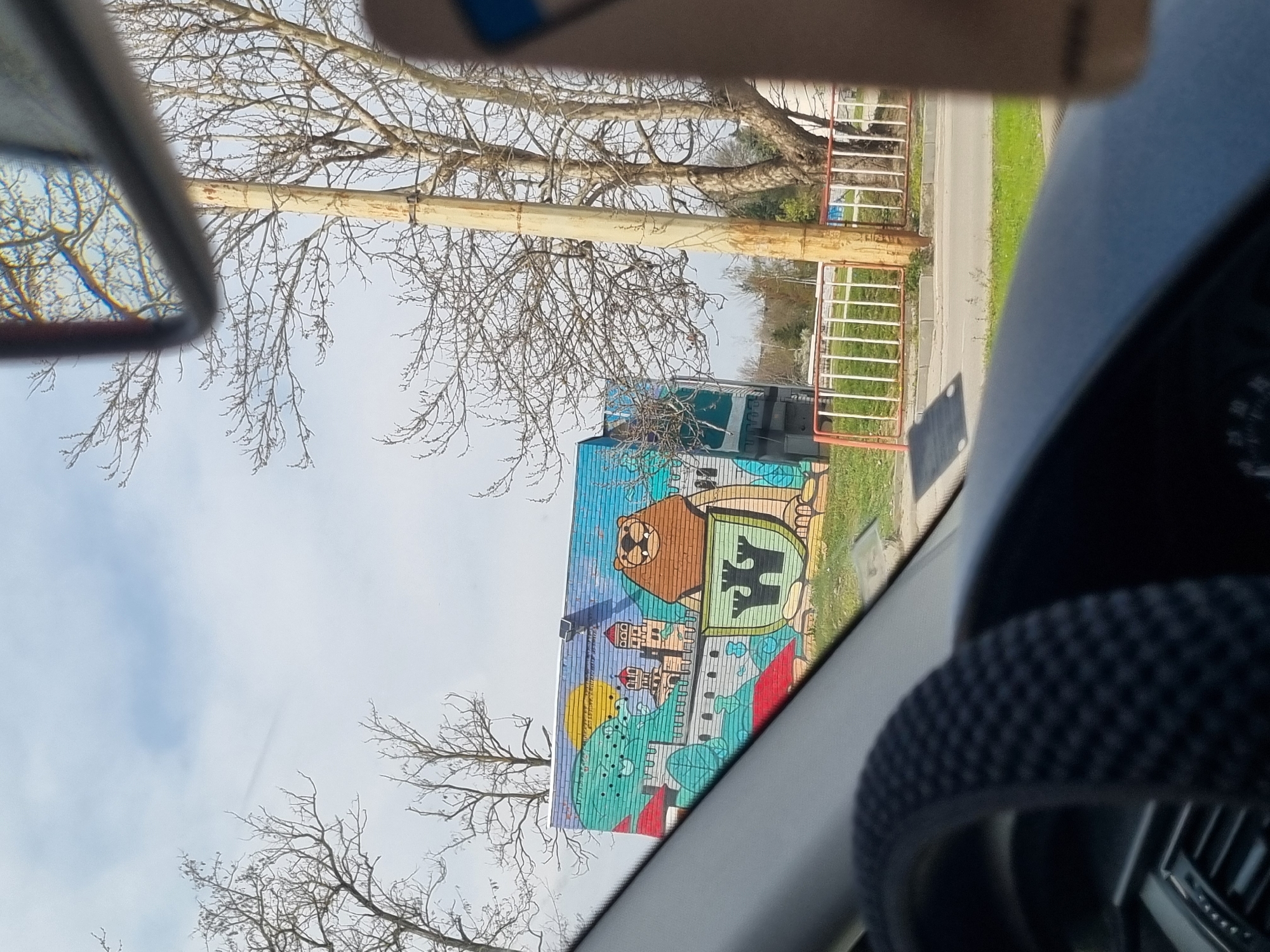 The image, taken from inside a car, shows a building with a large, colorful mural in Veliko Tarnovo, Bulgaria. The mural features a lion above a coat of arms with a black double-headed eagle, all on a light green background. Below this are stylized buildings and other shapes, rendered in light blue, yellow, and red. The building is partially obscured by trees with bare branches. Beyond the building, a light-colored fence, a road, and green grass can be seen. Parts of the car's interior, including the rearview mirror and steering wheel, are visible in the foreground. The sky is light blue with wisps of clouds.