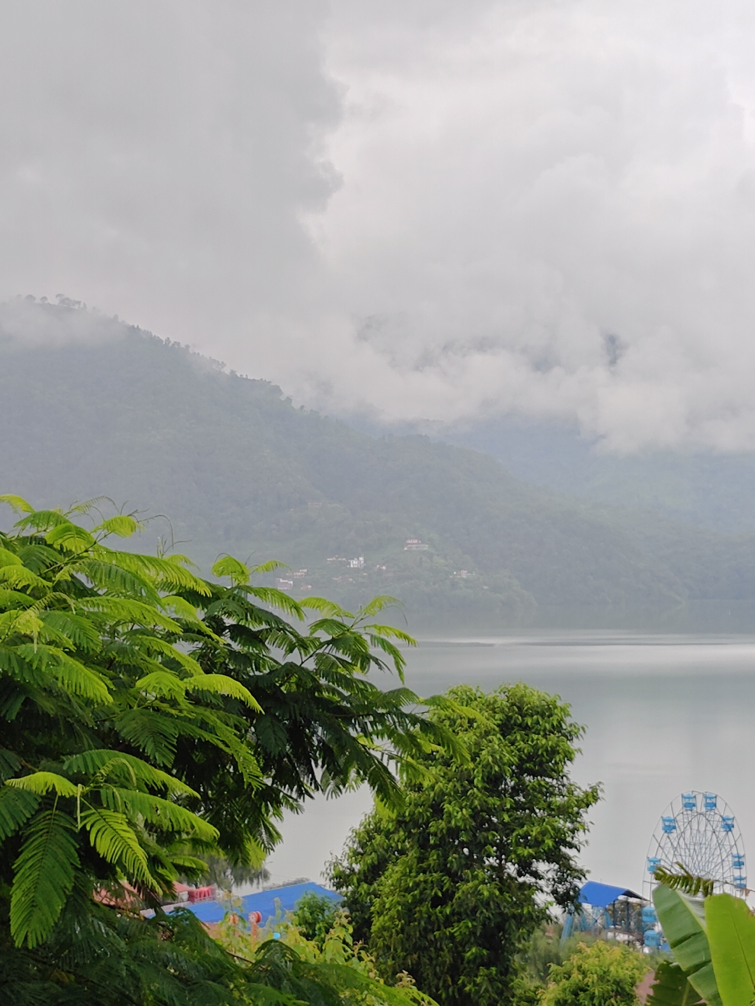 The image captures a scenic view of Phewa Lake in Pokhara, Nepal, under a cloudy sky. In the foreground, lush green foliage from trees partially obscures the view. To the right, a Ferris wheel with blue gondolas is visible, suggesting a recreational area. The lake itself is calm, reflecting the overcast sky. In the background, rolling hills covered in dense vegetation rise up, with a few small structures dotting the landscape. The overall atmosphere is serene and slightly muted due to the cloudy weather.