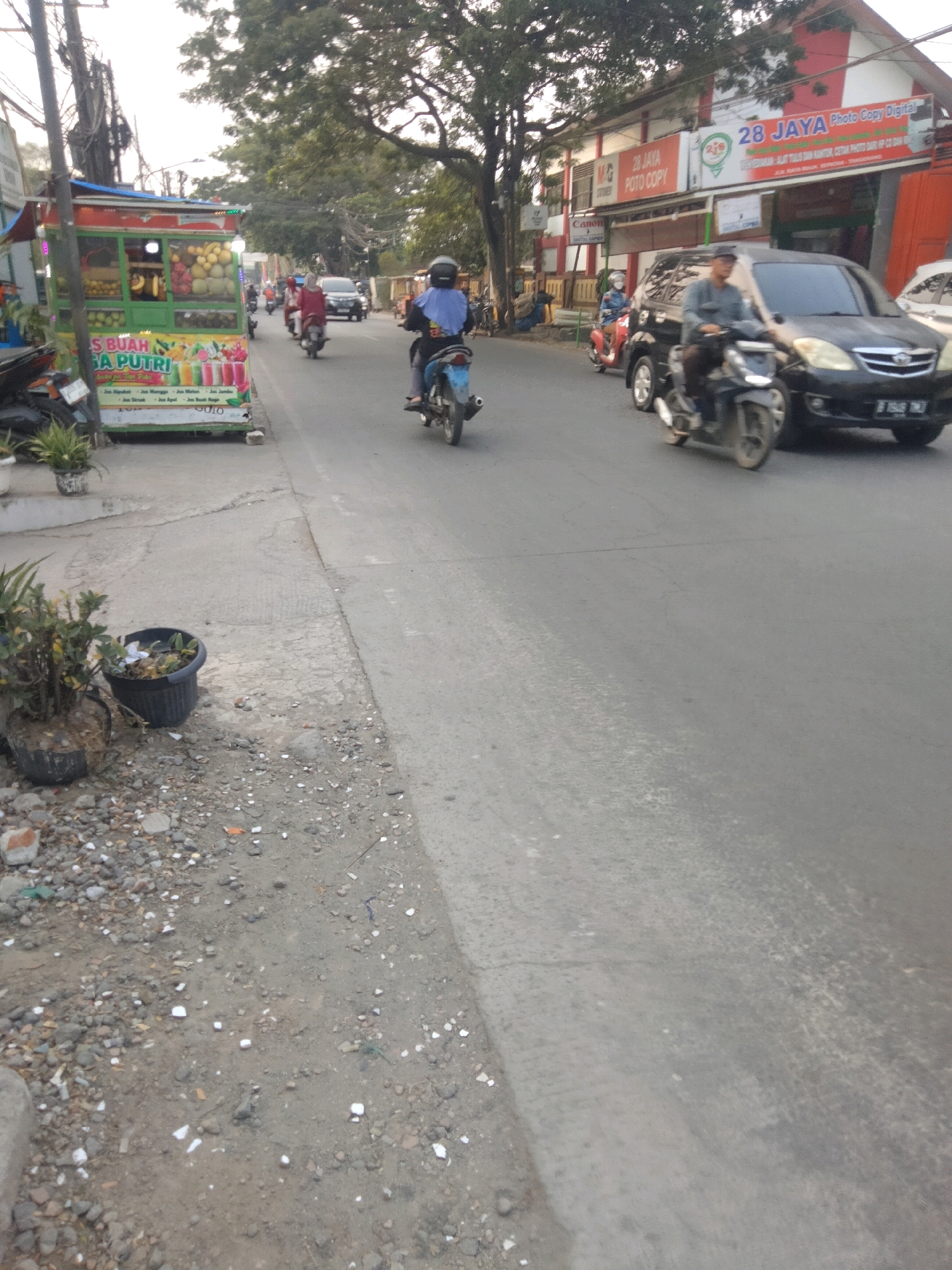 The image captures a street scene in Sepatan, Indonesia, likely during the day, given the lighting conditions. In the foreground, to the left, a mobile fruit juice stand is visible, displaying various fruits and colorful beverages. Adjacent to it, potted plants are placed on the side of the road, with some debris scattered on the ground.
The street itself is paved and appears to be a two-way road. Several motorcycles are present, with one rider wearing a blue headscarf and another in the background. A black car is in motion, and other vehicles are also visible further down the road. Buildings line the right side of the street, including a red and white building with signage for "28 JAYA Photo Copy Digital". Trees with dense foliage are also a prominent feature of the environment. The overall impression is of a bustling urban street with traffic and commercial activity.