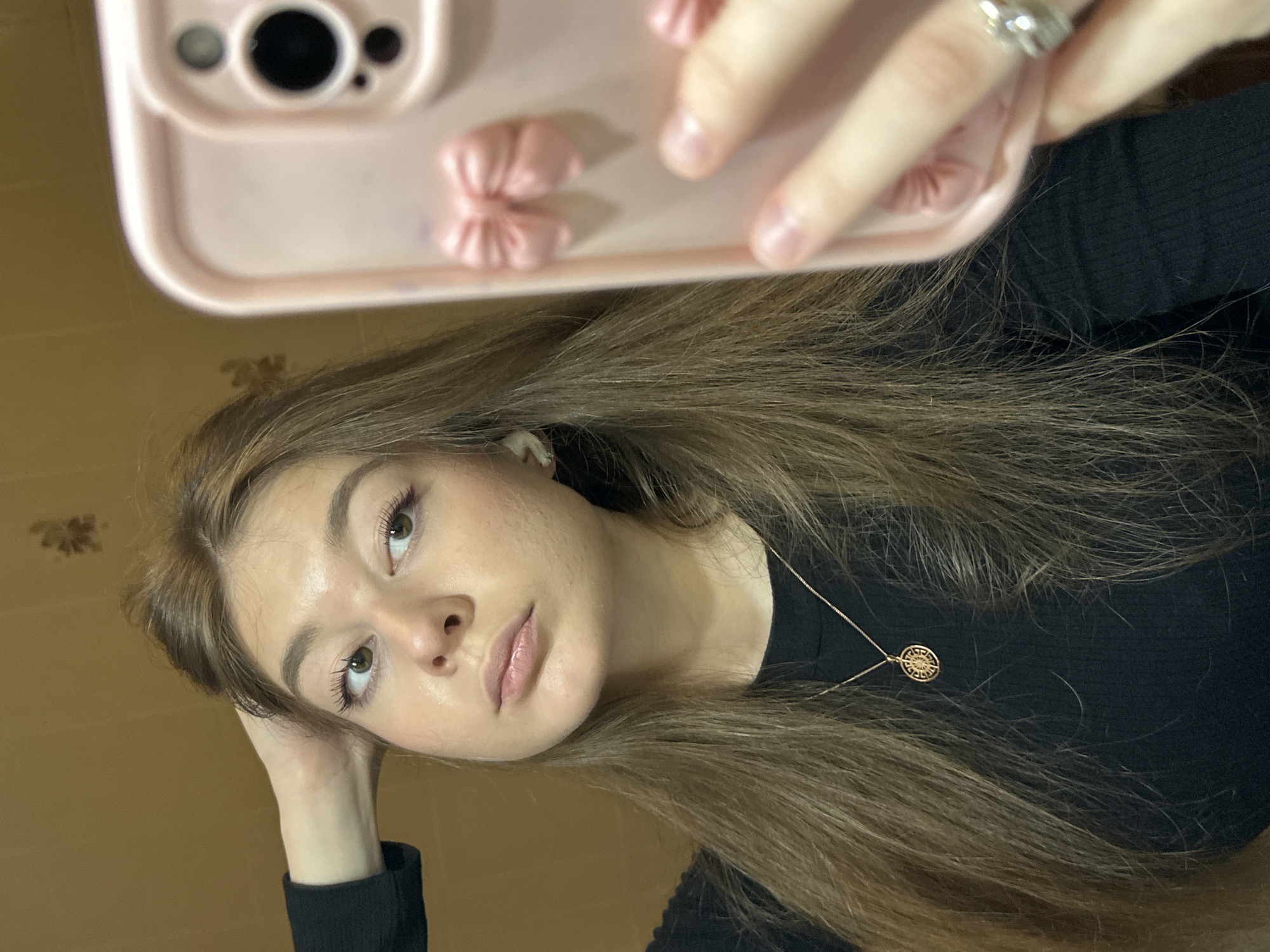 A young woman, appearing to be in Kazan, Russia, is captured in a selfie taken in what appears to be a mirror or highly reflective surface. The image shows her head and upper torso, tilted slightly. She has medium-long, light brown hair, which falls over her shoulders and chest. She is wearing a black long-sleeved top or sweater. Her face is turned towards the camera, with a neutral expression and slightly parted lips. Her eyes are light-colored, possibly green or hazel, and she has visible mascara on her lashes. Her left hand is bent, resting lightly against her jawline.
A gold necklace with a round, intricately designed pendant (featuring a geometric or sun-like pattern) is visible around her neck, resting on her black top.
In the upper part of the reflection, a pink smartphone with a distinct pink case is prominently displayed. The case features several small, decorative pink bows or ribbons on its back. The phone's camera lenses are visible. A hand, adorned with a silver ring on one finger, is holding the phone.
The background is a plain, light brown or tan wall, with a subtle, stylized floral or geometric pattern visible in the upper left corner of the scene. The lighting suggests an indoor setting, likely well-lit by natural or artificial light. There is no visible text in the image.