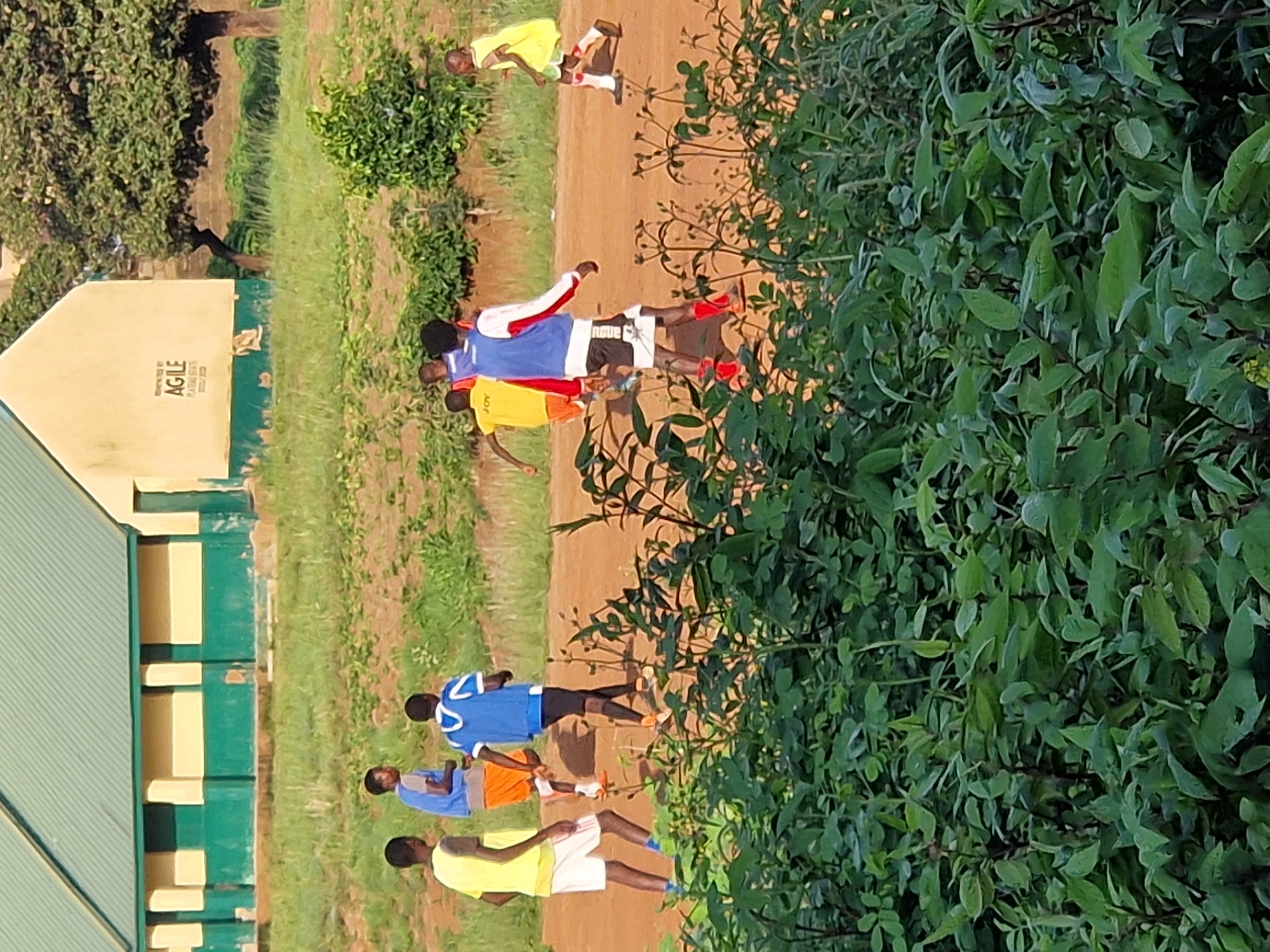 The image shows a group of young people playing soccer on a dirt field in front of a light-colored building with a green and yellow facade in Kabong, Nigeria. The building has a green metal roof and windows. The field is surrounded by dry grass and some leafy green bushes in the foreground. Several individuals are visible, wearing athletic attire. One person in a yellow shirt and white shorts is running. Another person in a blue shirt and orange shorts is also running. A third person in a blue jersey with white trim and black shorts is walking. A fourth person in a yellow shirt and green shorts is running towards the right. Another person in a yellow shirt is running on the right side of the frame. The overall atmosphere suggests a casual game of soccer in an outdoor educational or community setting.