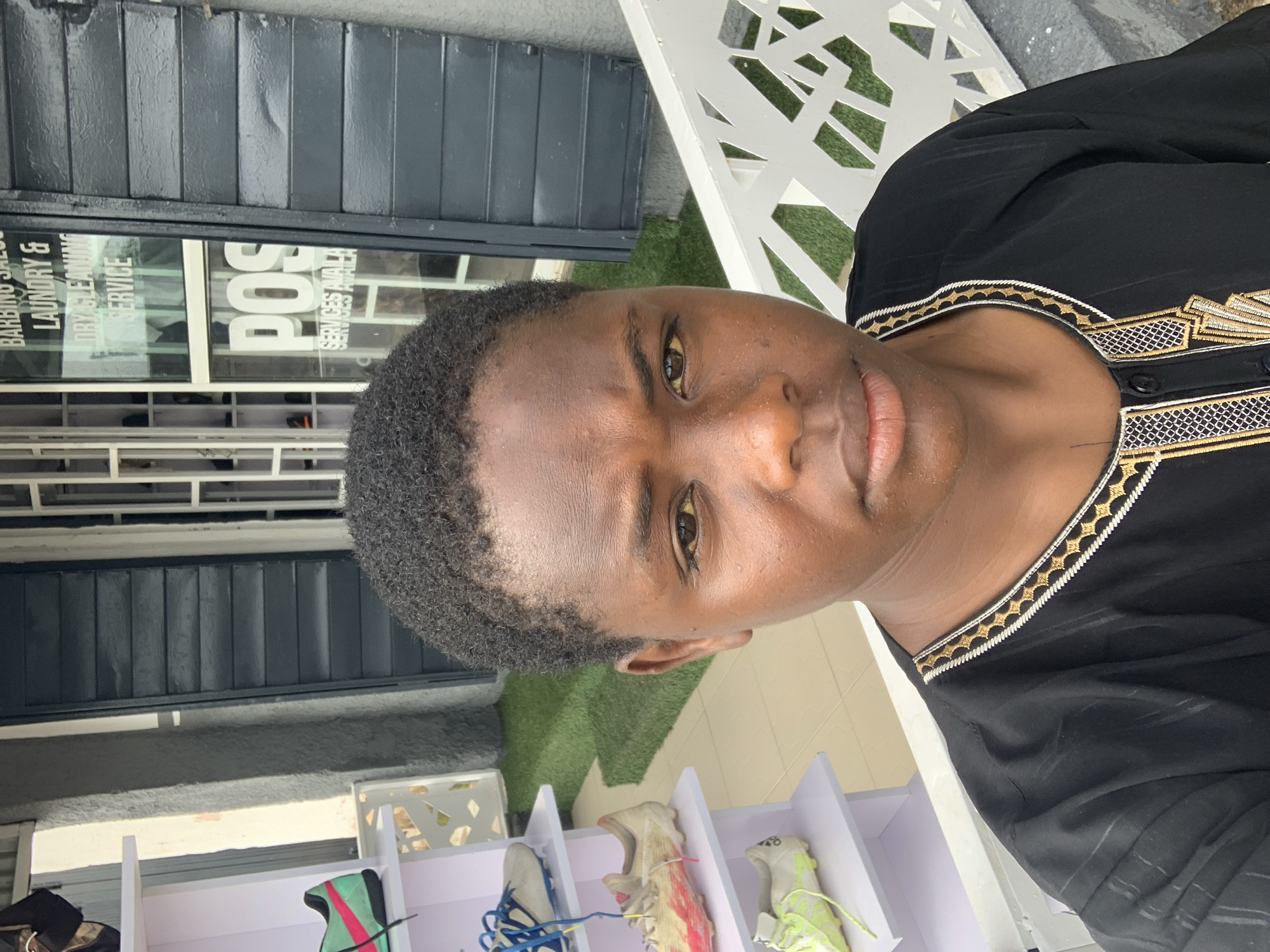 The image shows a close-up, portrait-style selfie of a young person. The subject has dark skin, short, dark curly hair, and is looking towards the camera with a neutral expression. They are wearing a black garment with ornate gold and white embroidery around the neckline. The background suggests an outdoor setting, possibly a shopfront.
Behind the subject, on the left, is a dark, ribbed surface, possibly a shutter or a wall. To the right of that is a glass window with text visible on it. The text appears to be in English and mentions "BARBING SALON," "LAUNDRY & DRY CLEANING SERVICES," and "POS SERVICES AVAILABLE." A large white sign with the letters "POS" in bold is also visible through the window.
Further to the right, a white latticed structure, possibly a chair or a bench, is visible against a backdrop of artificial turf. In the lower part of the image, a white shelving unit displays several pairs of athletic shoes. The lighting suggests it is daytime, and the overall atmosphere appears casual. The subject's facial features are clear, with prominent eyes and lips. The composition is a vertical shot, with the subject's head dominating the upper portion of the frame. The location context is Jos, Nigeria.