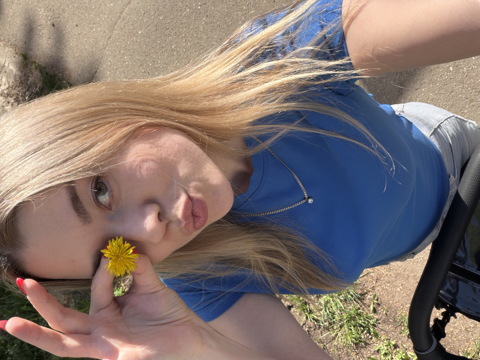 A young woman poses outdoors, holding a yellow flower close to the camera. She is fair-skinned with blonde hair, a light blue t-shirt, and a delicate necklace. Her fingers have red nail polish. She is making a kissing face at the camera. A stroller handle is visible on the right side of the image. The location appears to be an outdoor area with grass and pavement. This photo was taken in Chaikovsky, Russia.
