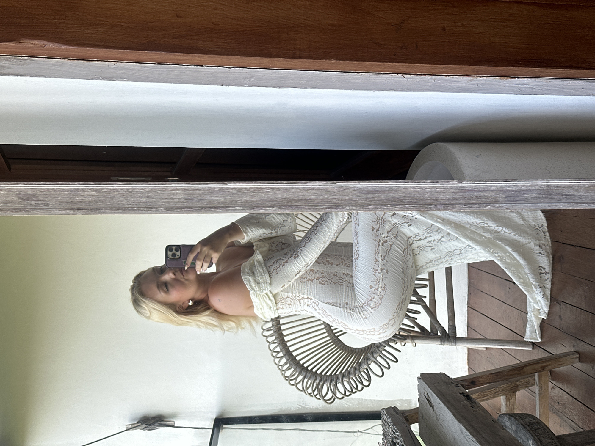 A young, blonde woman is seated in a woven chair, holding a smartphone to take a selfie in a mirror. She is wearing a long, off-the-shoulder, white lace dress. The scene appears to be an outdoor balcony or patio with a wooden floor and railing. To the left of the woman, there is a rustic wooden stool. To the right of the woman, a doorway with a dark wooden frame is visible, leading into a darker interior space. The woman's reflection is captured in a large mirror to her right. The location is identified as North Kuta, Indonesia, likely a private residence or hotel.