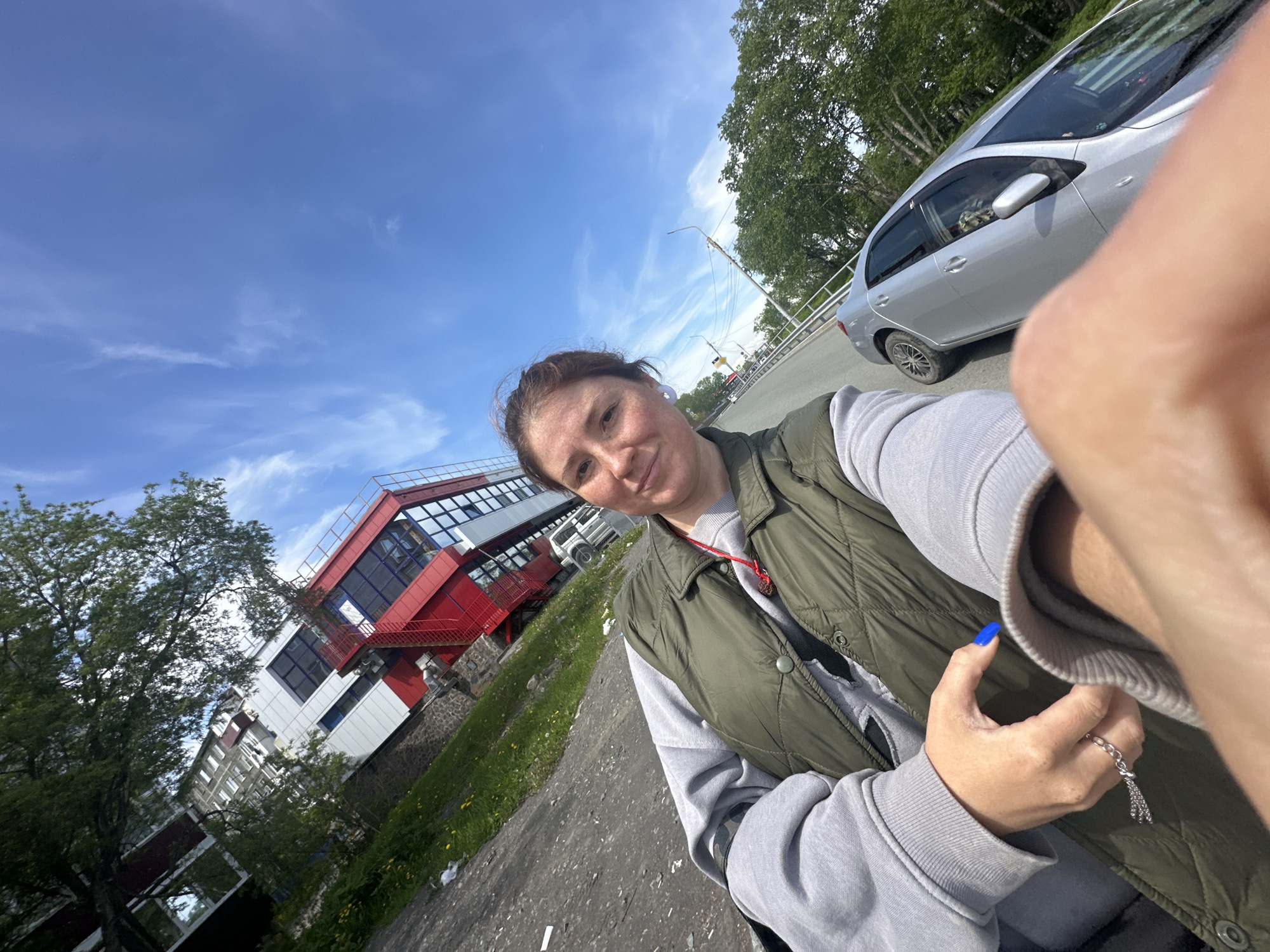 A woman with auburn hair pulled back, wearing a grey hoodie and a green vest, takes a selfie outdoors. She has freckles on her face and is wearing a white earring in her left ear and a silver ring with tassels on her right hand. Her fingernails are painted bright blue. Behind her, a modern building with white, red, and grey sections and prominent red external stairs is visible. The sky is bright blue with wispy white clouds. A silver car is parked on the right side of the road. The scene appears to be in an urban or suburban area with a road, trees, and a grassy embankment. The presence of the building and car, along with the general atmosphere, suggests a location in Petropavlovsk-Kamchatsky, Russia.