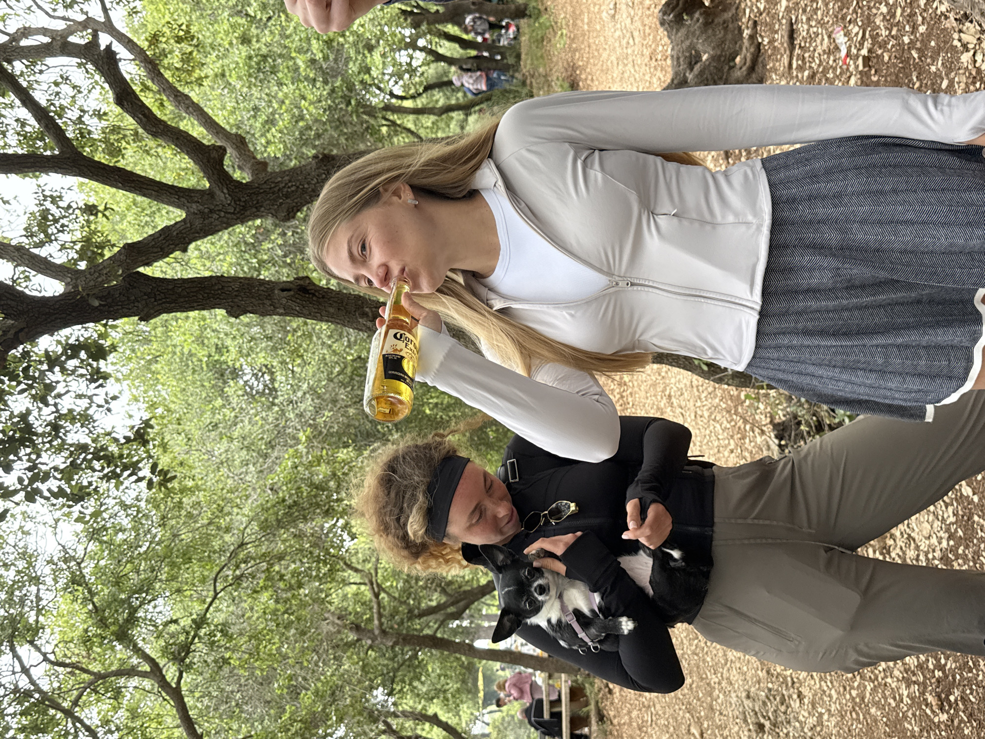 The photograph, taken outdoors in a wooded area, depicts two women. The woman in the upper portion of the frame is blonde and wearing a light-colored zip-up jacket, a white undershirt, and a dark herringbone skirt. She is drinking from a bottle of Corona beer. The woman in the lower portion of the frame is wearing a black long-sleeved shirt, dark pants and a black headband. She is holding a small black and white dog. The surroundings are characterized by trees with green foliage and a ground covered in leaves. Other individuals are indistinctly visible in the background. Although asked to include information from Nesher, Israel, there is no identifiable information in the image to locate the scene in that city.