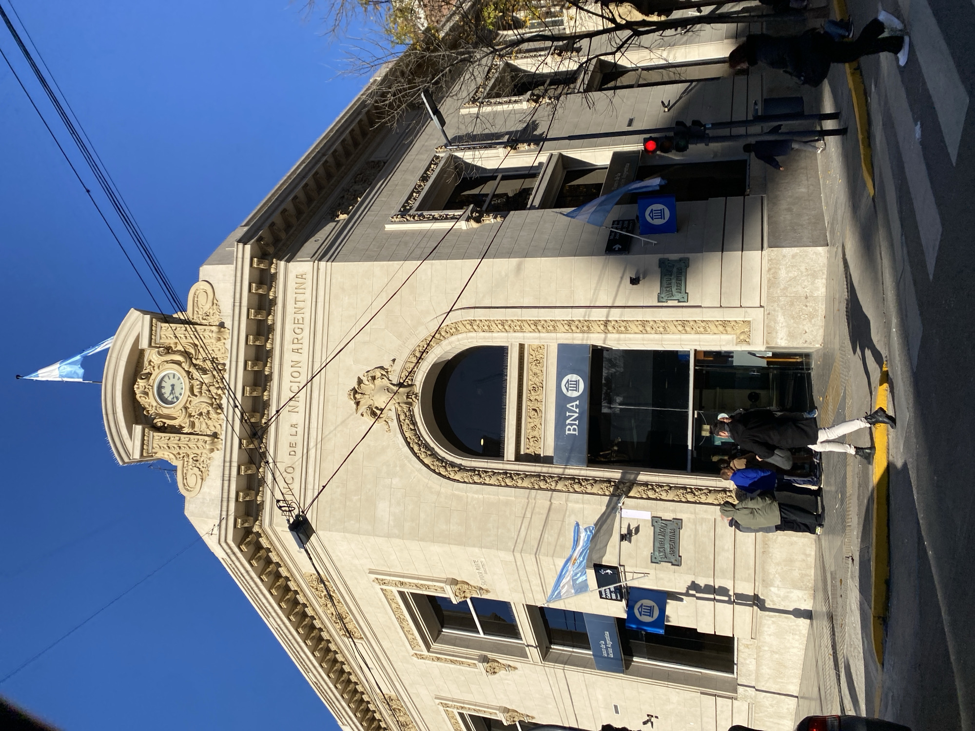 The image depicts the Banco de la Nacion Argentina building in Buenos Aires, Argentina. The building's facade is visible, featuring ornate architectural details, including a clock tower and arched windows. Signage and flags of Argentina are displayed on the building. Pedestrians are present on the sidewalk in front of the building, and a crosswalk is visible. A street and traffic lights are also visible.