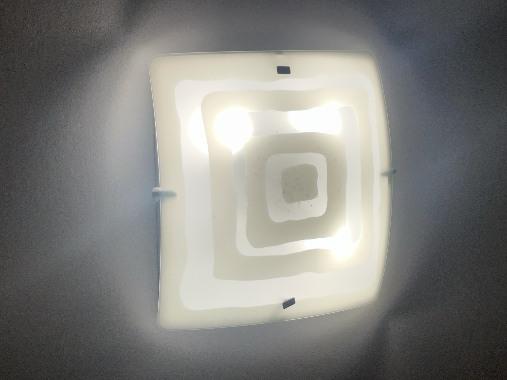 An overhead view captures a square, flush-mount light fixture mounted on a light gray, textured ceiling or upper wall in Novi Sad, Serbia. The fixture features a milky white, opaque diffuser with slightly rounded corners. Visible through the diffuser are two bright, round light sources, predominantly in the upper half, which illuminate the fixture and the surrounding surface.
The diffuser itself has a distinctive design, with a layered, concentric square pattern subtly etched or molded into its surface, creating a decorative effect. Small, dark, rectangular clips are visible on the top and bottom edges, holding the diffuser in place. Several tiny dark specks, possibly dust, are noticeable on the central part of the diffuser.
The light emanating from the fixture creates a bright glow directly around it, highlighting the subtle texture of the ceiling. This glow gradually fades into the darker, uniform gray of the wider ceiling area, suggesting the light is the primary source of illumination in the space. No other objects, people, or text are visible in the image. The scene appears to be an interior space, with the artificial light indicating either evening or an interior room without natural light.