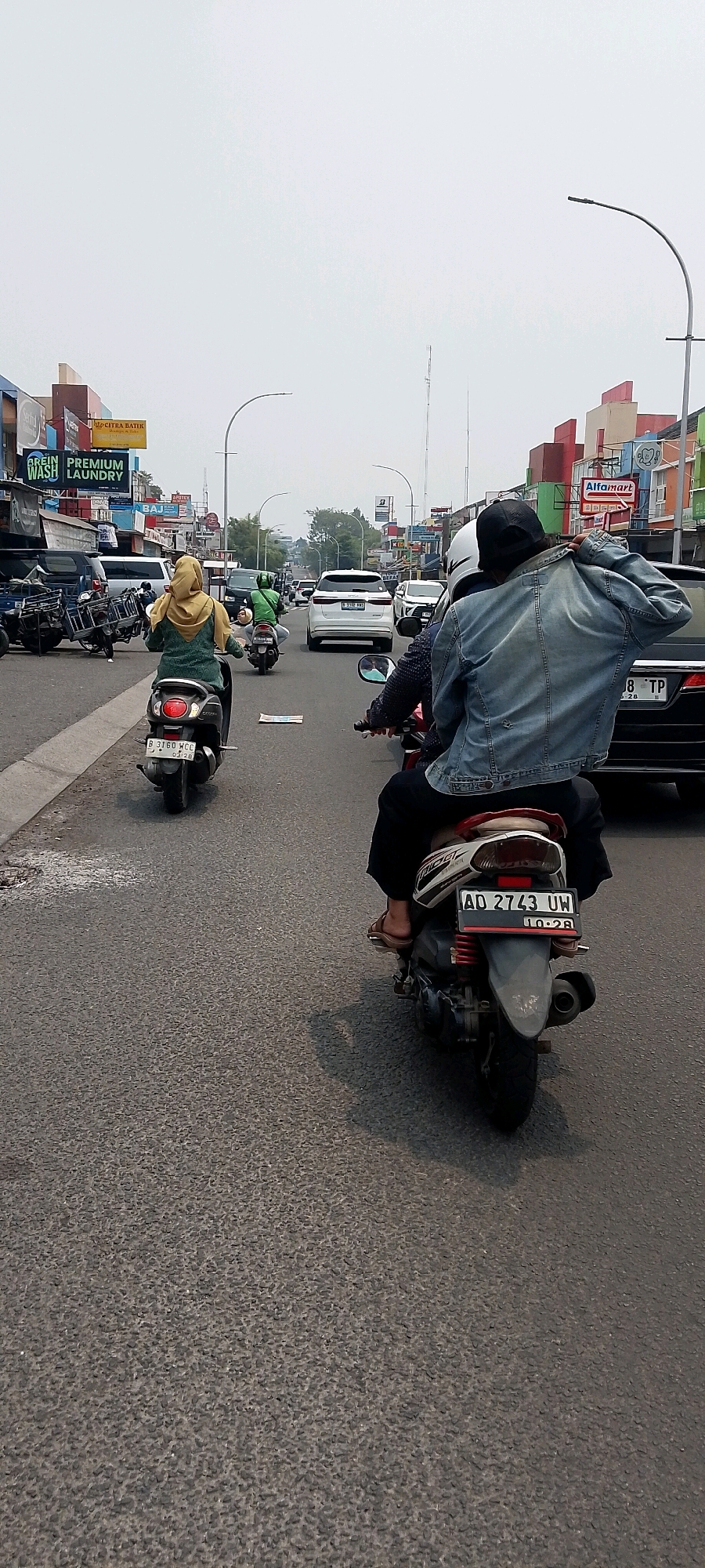 The image shows a street scene in Cileungsi, Indonesia, characterized by a moderate amount of traffic, including motorcycles and cars. The perspective is from a vehicle traveling on the road, looking forward and slightly to the right. In the foreground, two people are riding a motorcycle. The person in the back is wearing a denim jacket and a black cap, with their back to the camera, and appears to be adjusting their jacket. The person in the front is wearing a helmet. The motorcycle's license plate is visible and reads "AD 2743 UW".
Further ahead on the road, another motorcycle with a rider wearing a yellow hijab and a green top is visible. Several cars are also present in the traffic, including a white SUV with the license plate "B 3358 FKD". The buildings lining the street are a mix of commercial establishments, some with prominent signage, including "Alfamart", "APOTEK", and "PREMIUM LAUNDRY". The sky is overcast. The road surface is asphalt. The overall atmosphere suggests a busy urban environment.