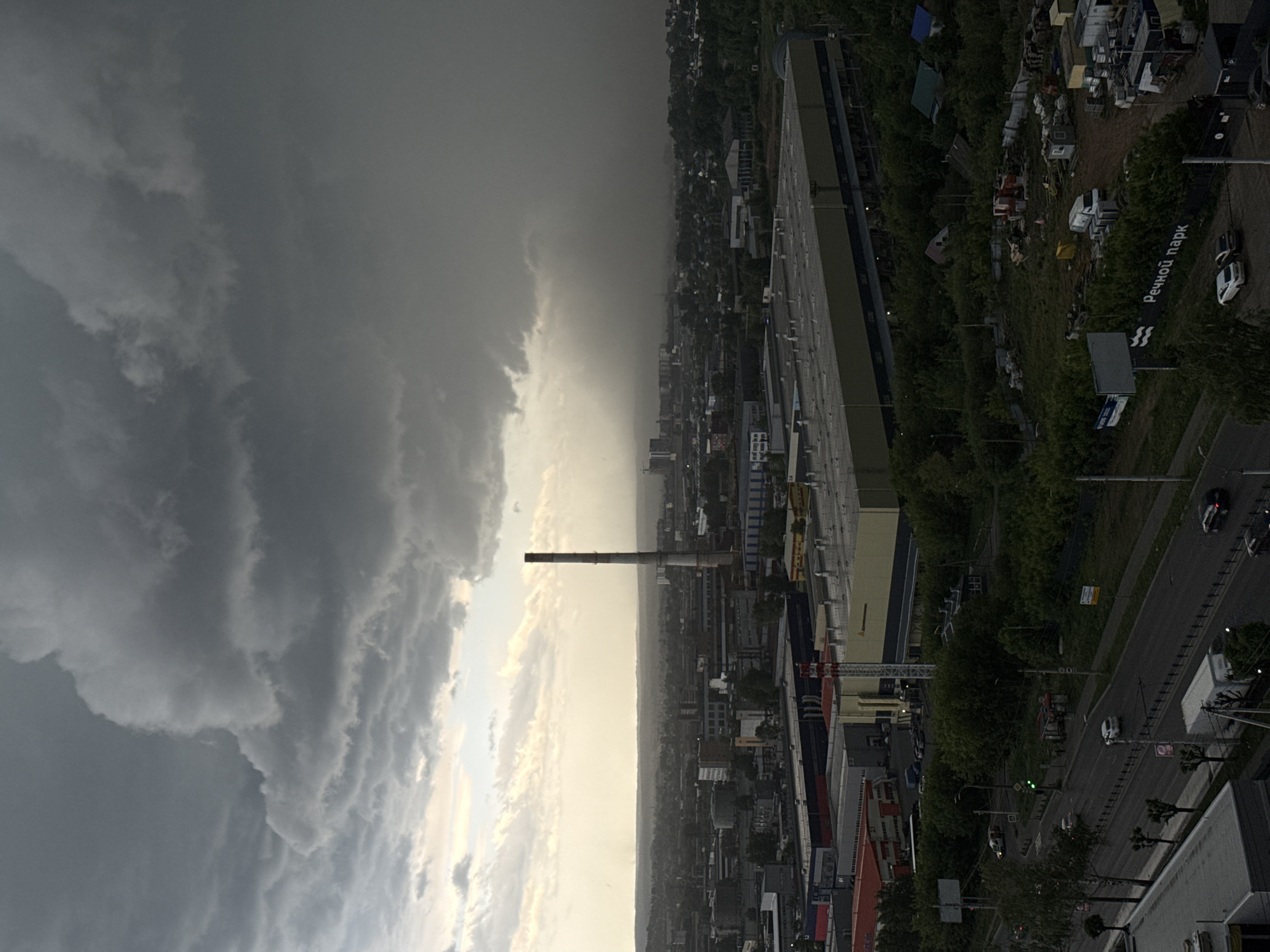 The image depicts an urban landscape under a stormy sky, centered on a tall industrial chimney. The location is Izhevsk, Russia, discernible from the sign "Речной парк" (River Park) on the right side of the image.
The left side of the image is dominated by a dense, dark cloud formation, indicating a storm. The right side reveals an urban setting with buildings, roads, and trees. The central focus is the chimney, which rises vertically amid the buildings.
Roads lined with trees and streetlights are visible. There are cars on the road, suggesting urban activity. A large building with a light-colored roof is present. The background shows other urban structures and green areas.