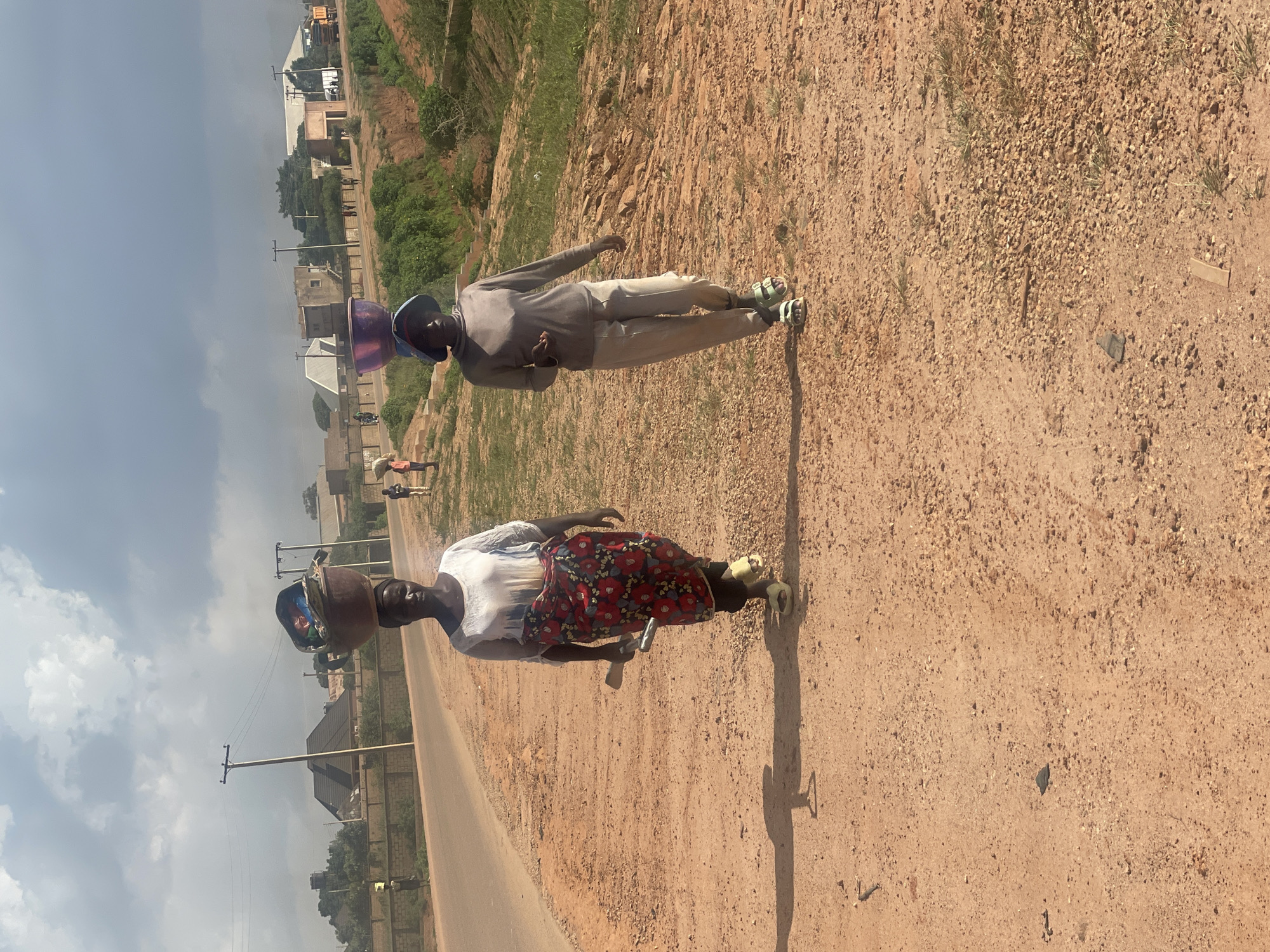 Two women walk along a dusty, unpaved road in Kwang, Nigeria. The woman on the left carries a large, brown basin filled with various items on her head, wearing a white top and a red and floral patterned skirt. The woman on the right wears a grey long-sleeved shirt and light-colored pants, with a blue hat and a purple basin balanced on her head. The environment is rural, with sparse vegetation, scattered buildings in the background, and utility poles lining the road. The sky is cloudy.