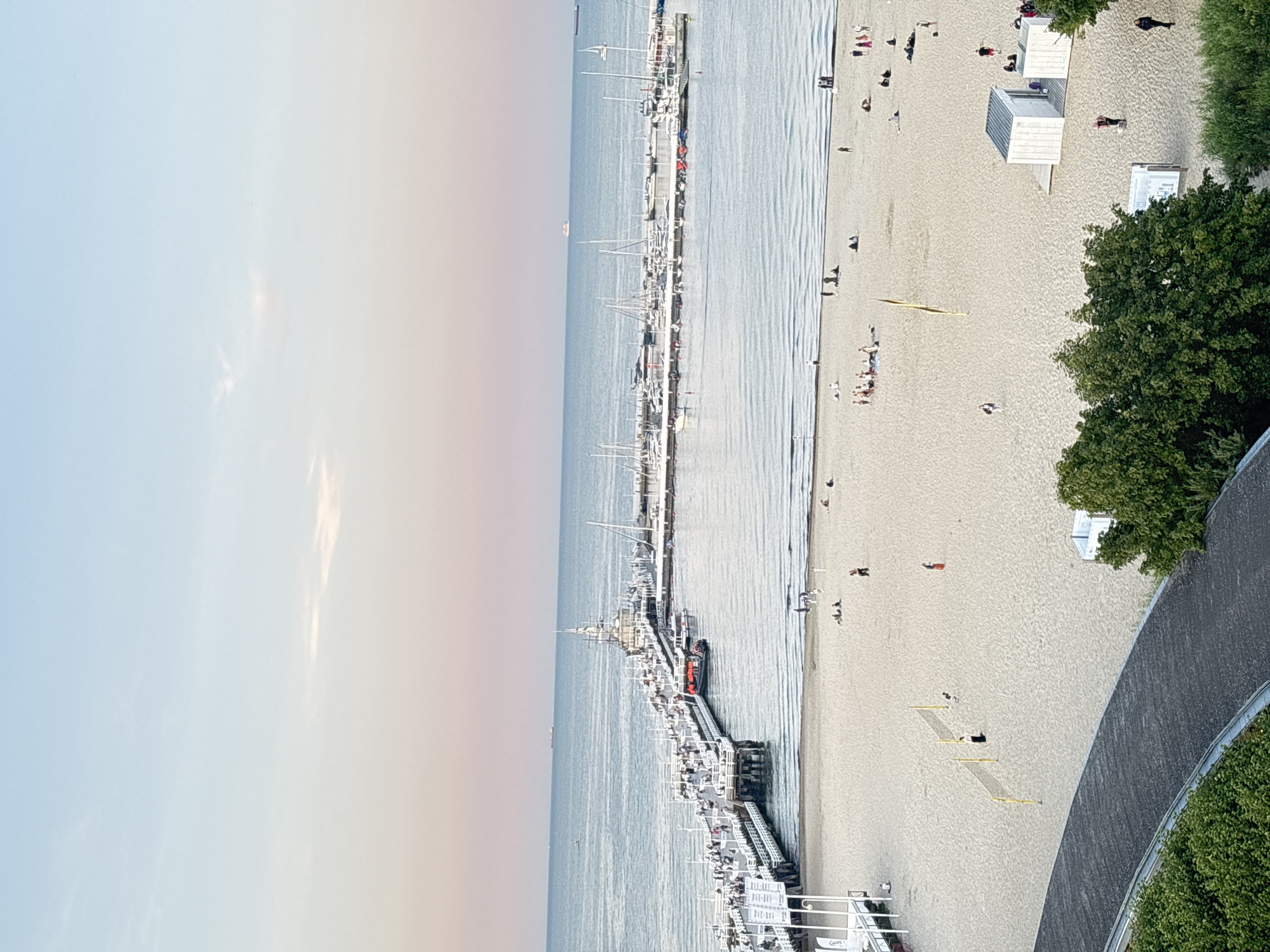 The image shows an aerial view of a beach and pier in Sopot, Poland. The sky is a gradient of soft pink and blue. The sea is calm with small waves. A long white pier extends from the beach into the water, lined with people and a lighthouse-like structure at its end. A marina with numerous sailboats and yachts is located to the right of the pier. The beach is sandy and populated with people sunbathing, walking, and playing volleyball. There are also several beach huts and some greenery along the edge of the beach. The overall scene depicts a pleasant summer day at a popular seaside resort.