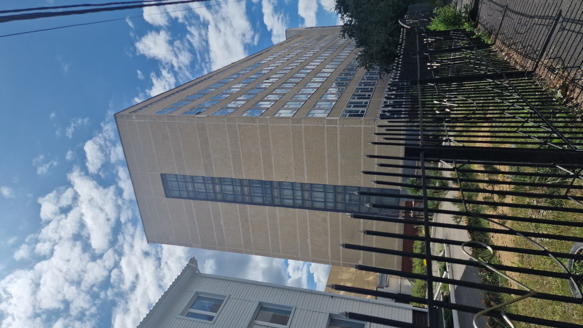 The image captures a large, light-colored brick building situated in an urban environment, likely in Kostanay, Kazakhstan. The building is several stories high, with a prominent vertical section of dark-tinted windows running up its center. Rows of windows are also visible along the length of the building's facade. To the left of this main building is a smaller, white-paneled structure with visible windows. In the foreground, a black metal fence with pointed finials runs across the scene, partially obscuring the lower portion of the buildings and the surrounding grounds, which appear to be a mix of grass, dirt, and a paved walkway casting shadows. The sky above is blue with scattered white clouds, suggesting daytime. No people are visible in the image. The presence of utility wires is also noted in the upper right corner of the frame.