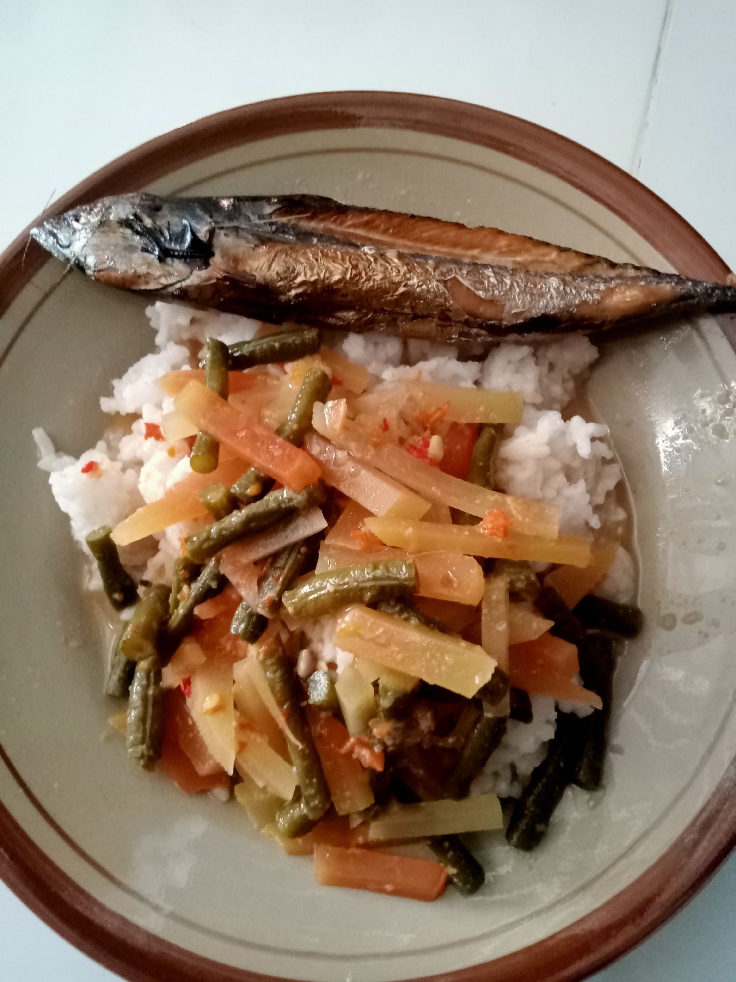 A close-up, top-down shot shows a plate of food in Simo, Indonesia. The plate contains white rice topped with a medley of vegetables and a whole, cooked fish. The vegetables appear to be green beans and possibly chayote or papaya, cut into strips and mixed with red chili pieces and a light sauce. The fish, a small, silvery specimen, is positioned horizontally on top of the rice. The plate itself is ceramic with a brown rim. The background is plain and appears to be a white surface, possibly a table.