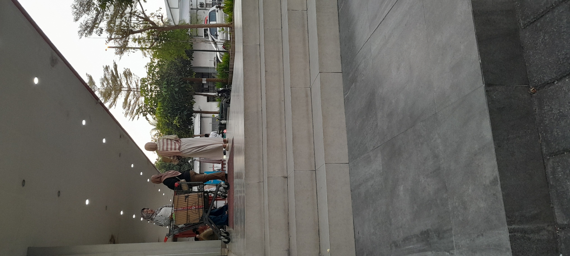 In the city of Rogojampi, Indonesia, at what appears to be the entrance to a commercial building, three individuals are visible on a raised platform. Two of the individuals, dressed in hijabs, are standing near a shopping cart. One is facing the camera, while the other is partially obscured. The third individual, also wearing a hijab and a striped top and long skirt, is facing away from the camera. Behind them, a line of trees and a white building with a fence are visible. A silver car and a parked motorcycle are also present in the background. The foreground shows a series of wide, concrete steps leading down to a tiled ground area. The lighting suggests it is either early morning or late afternoon.