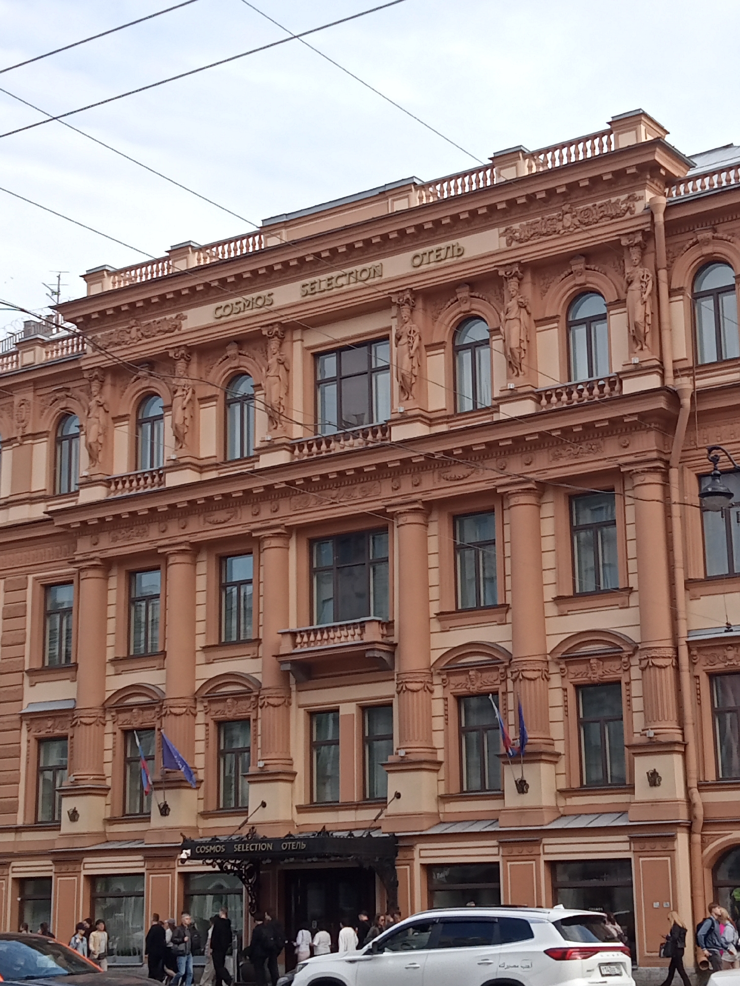 The image shows a facade of a multi-story building with ornate architectural details. The building is painted in a light brown or salmon color and features columns, decorative carvings, and arched windows. The text "COSMOS SELECTION ОТЕЛЬ" is visible above the main entrance. Several people are walking on the sidewalk in front of the building, and some cars are parked on the street. Two flags, one red, white, and blue, and another blue, are hanging from the building. The sky is overcast.