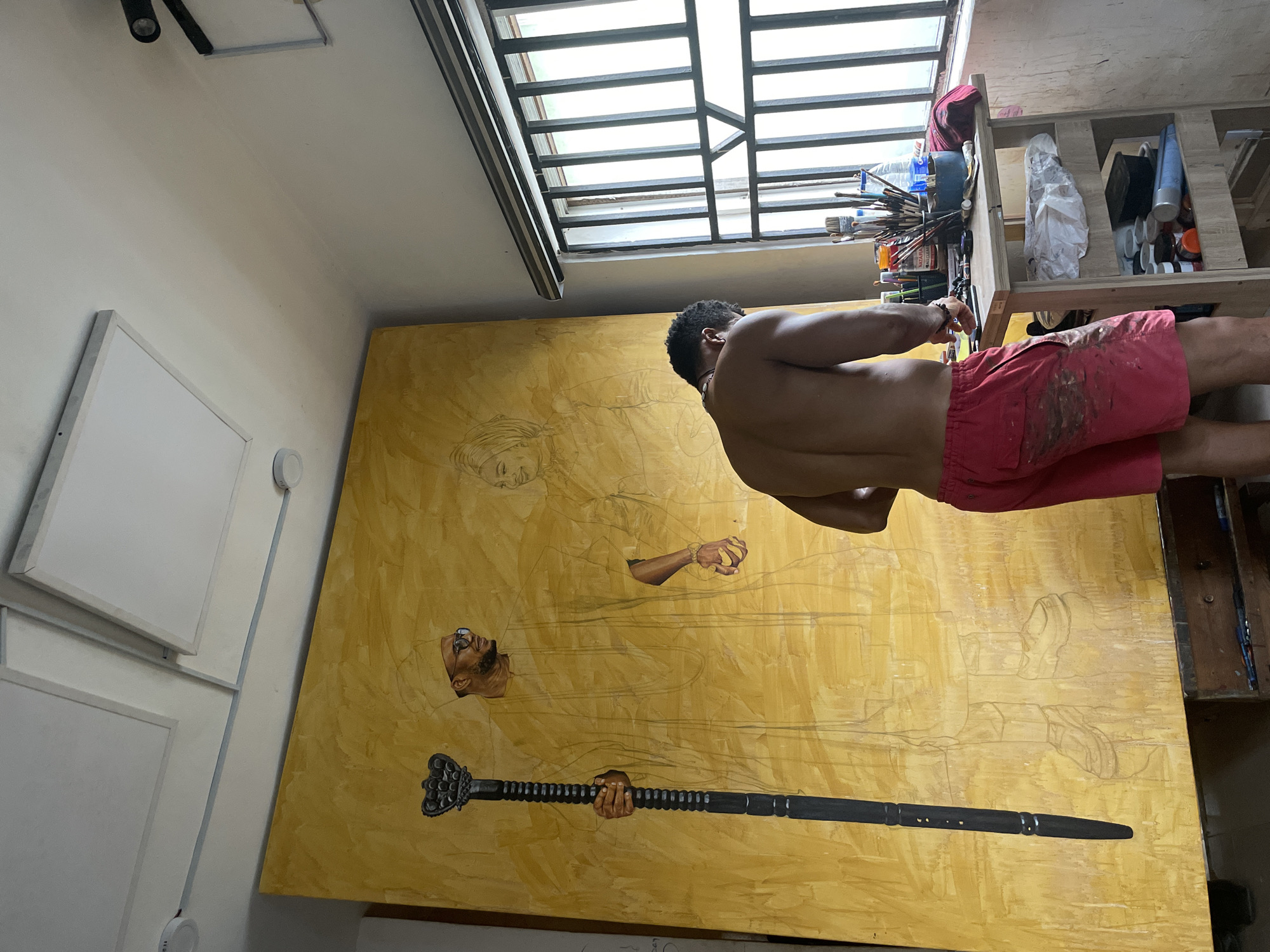 An artist is captured from behind, working intently on a large, predominantly yellow canvas in what appears to be an art studio in Ado, Nigeria. The shirtless man wears red shorts, stained with paint, and is positioned slightly to the right of the canvas, facing towards it.
The massive canvas features a detailed pencil sketch of multiple figures, overlaid on a warm yellow underpainting. One central figure, depicted with a beard and glasses, has his head and hands partially painted in, with one hand grasping a distinctive black staff-like object. Above this, another pair of hands—one clenched, the other holding an indistinct item—is sketched. To the left, the outline of a head wearing a headwrap is visible. Other figures are faintly sketched into the background, suggesting a complex narrative composition.
The studio environment is brightly lit by natural light streaming through a large, gridded window positioned on the ceiling or upper wall. To the right, a wooden shelf or cart holds a variety of art supplies, including numerous paintbrushes, tubes of paint, and bottles. Visible text on some containers includes "NATARA" and "ALCHEMIX." The walls are white, with several white-framed panels, possibly blank canvases or light fixtures, mounted on the left side. The scene conveys a focused and productive atmosphere within a creative workspace during daytime.