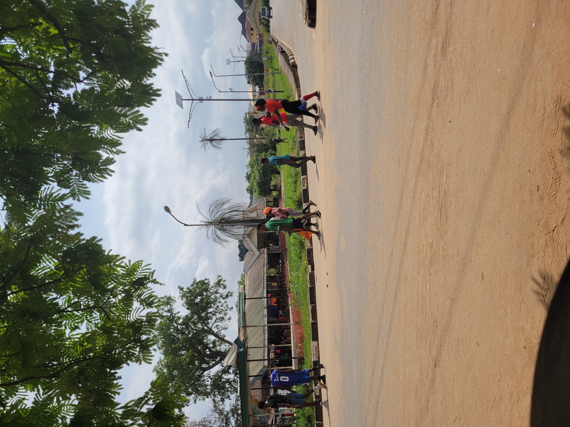 In Egbagu, Nigeria, on a bright, partially cloudy day, a group of six people is crossing a wide, dusty road. The individuals, varying in age and gender, are walking from left to right across the frame. One person, wearing a blue basketball jersey with the number 0, is in the foreground on the left, followed by another person in a black shirt and blue shorts. A person in a green shirt and dark shorts, holding an orange bag, is further ahead on the right. Behind them, three more individuals are walking, one in a red shirt and black pants, another in an orange top and black leggings, and a third person in a blue shirt.
To the left of the road, a long, open-sided structure with a corrugated metal roof, likely a market or seating area, is visible. Several patrons are seated inside. Green grass and trees line the area behind the structure, and palm trees are scattered throughout the background. In the distance, there are houses and other buildings. The road itself is paved, with a sandy shoulder on the left and a concrete curb on the right. Several streetlights, some with solar panels, are present along the road. The image is taken from a low angle, with leafy branches from a tree overhead casting shadows in the upper part of the frame.