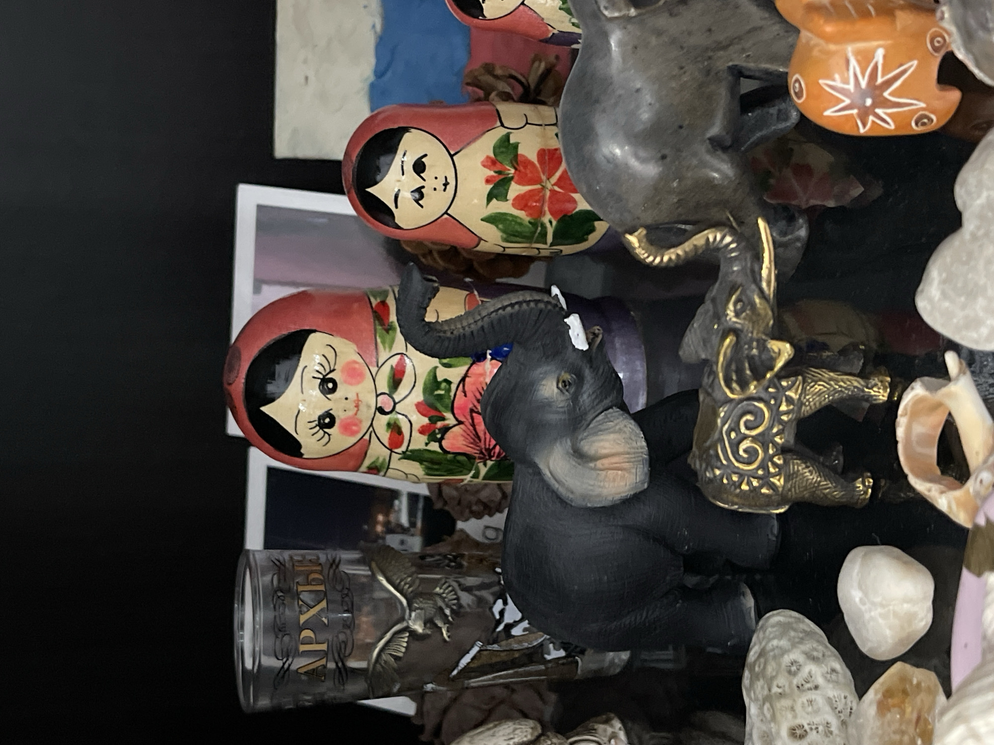 The image shows a collection of objects on a reflective black surface. There are two Russian nesting dolls (matryoshka dolls) with floral designs and red headscarves, a gray elephant figurine, a small golden elephant figurine, a glass with the inscription "АРХЫЗ" (Arkhyz), possibly a souvenir from Arkhyz, a resort town in the Karachay-Cherkess Republic, not Stavropol. There are also various stones and shells scattered around. The presence of the "АРХЫЗ" glass does not indicate the exact location, and the objects appear to be souvenirs or decorative items.