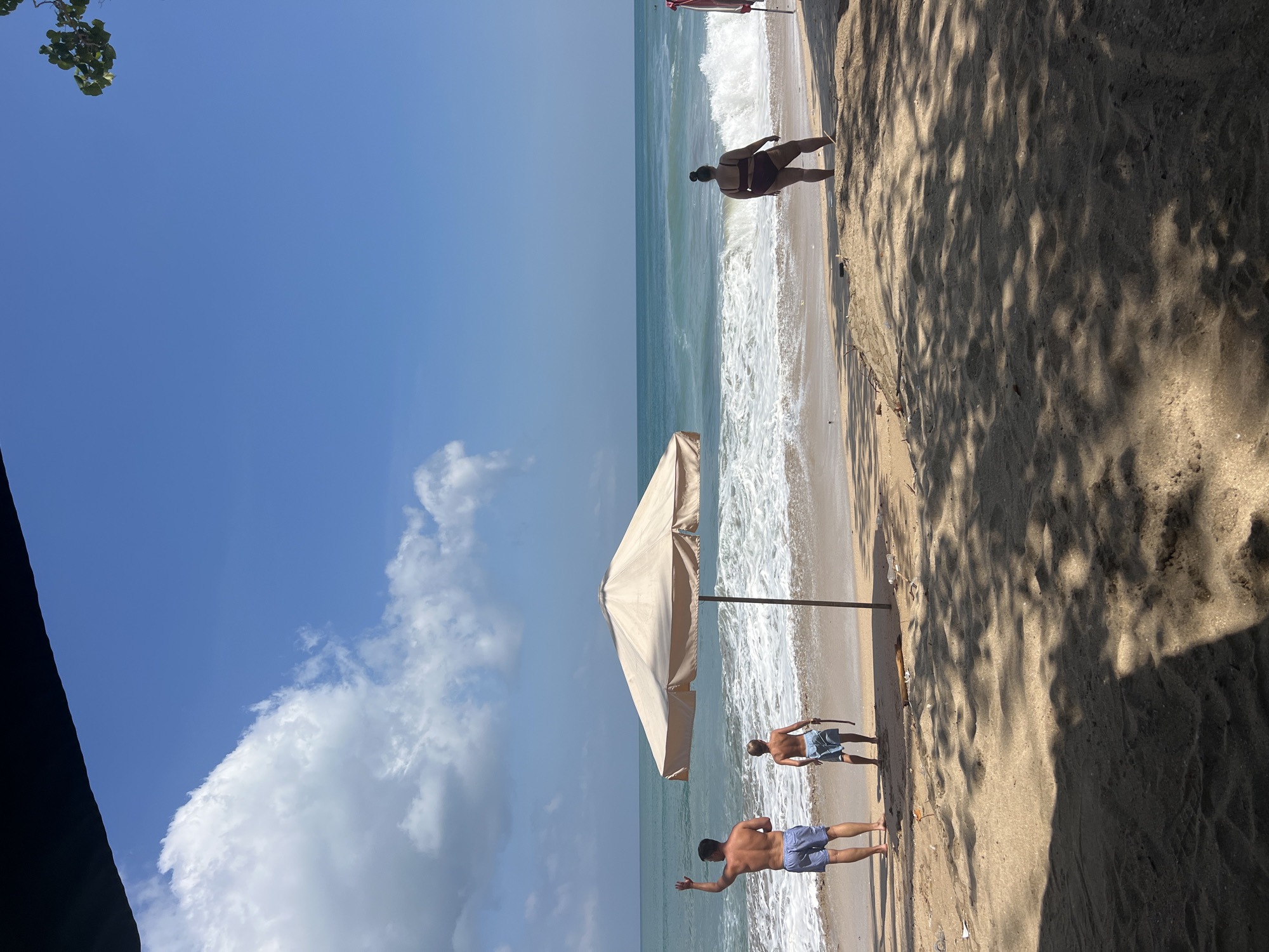 A beach scene unfolds under a bright blue sky with scattered white clouds. The sand, a light tan color, is textured with ripples and shaded by dappled sunlight filtering through unseen foliage. The ocean meets the shore in a series of white-capped waves, with the water transitioning from a pale turquoise near the beach to a deeper blue on the horizon.
In the foreground, partially visible on the left, is the edge of a dark object, possibly a structure or awning. Further into the scene, a large, light-colored beach umbrella stands tall on the sand, its canopy casting a shadow.
Several people are present on the beach. Near the water's edge, a person in a dark swimsuit faces away from the camera, with waves lapping at their feet. Further down the beach, two individuals in blue patterned swim shorts are walking near the water. One of them is facing the camera, gesturing with their left hand. Another person, further up the beach, is also visible.
The scene appears to be set in the daytime, likely morning or afternoon, given the bright sunlight and clear skies. The location is Kuta, Indonesia, a popular beach destination. The overall mood is relaxed and leisurely, suggesting a vacation or a day spent enjoying the seaside. There is no visible text in the image.