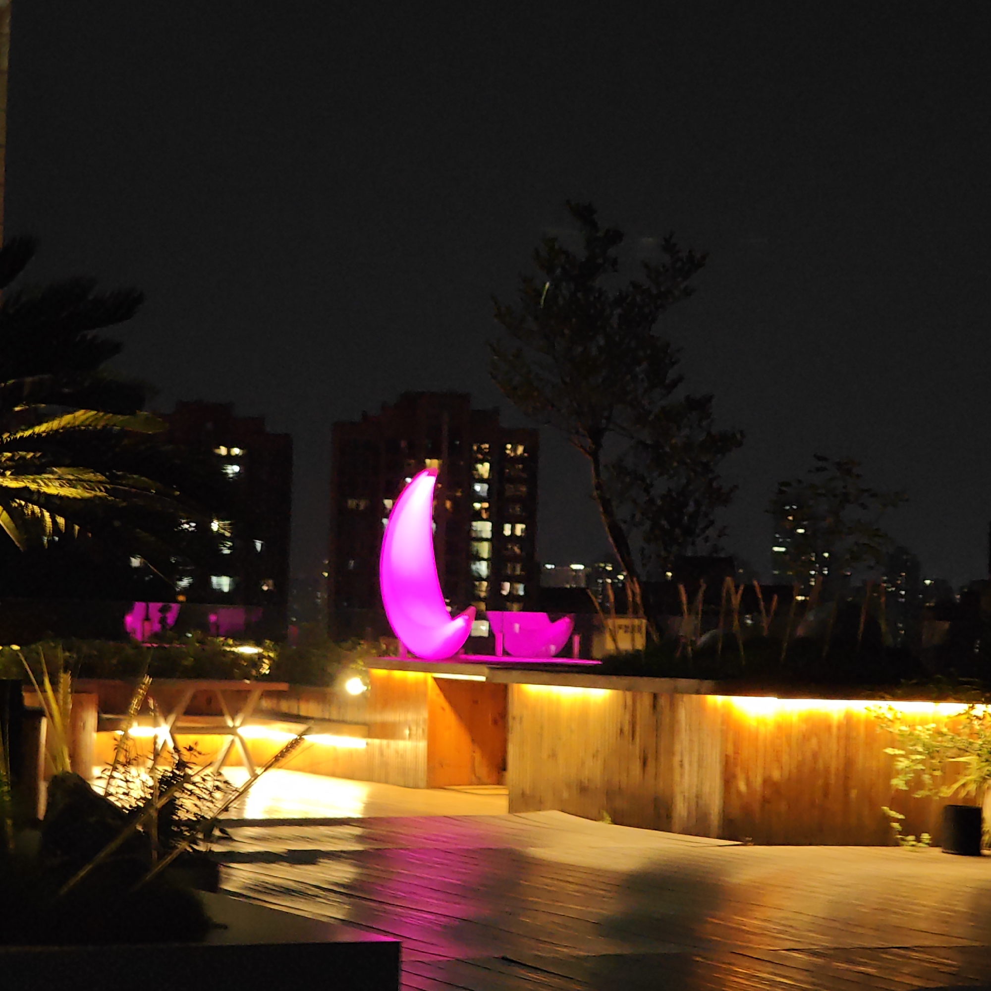 The image captures a nighttime scene in Yuzhong District, China, featuring a rooftop patio or deck. Dominating the view are two large, illuminated pink structures: one crescent-shaped and the other resembling a bowl or seating. Behind the structures, a building with illuminated windows indicates an urban setting. Below the pink structures, the deck surface is wooden, with warm yellow light emanating from beneath an elevated section. Palm trees and other greenery are interspersed throughout the deck area, adding a touch of nature to the urban rooftop. The overall atmosphere is modern and stylish, with the unusual lighting and design elements contributing to a unique ambiance.