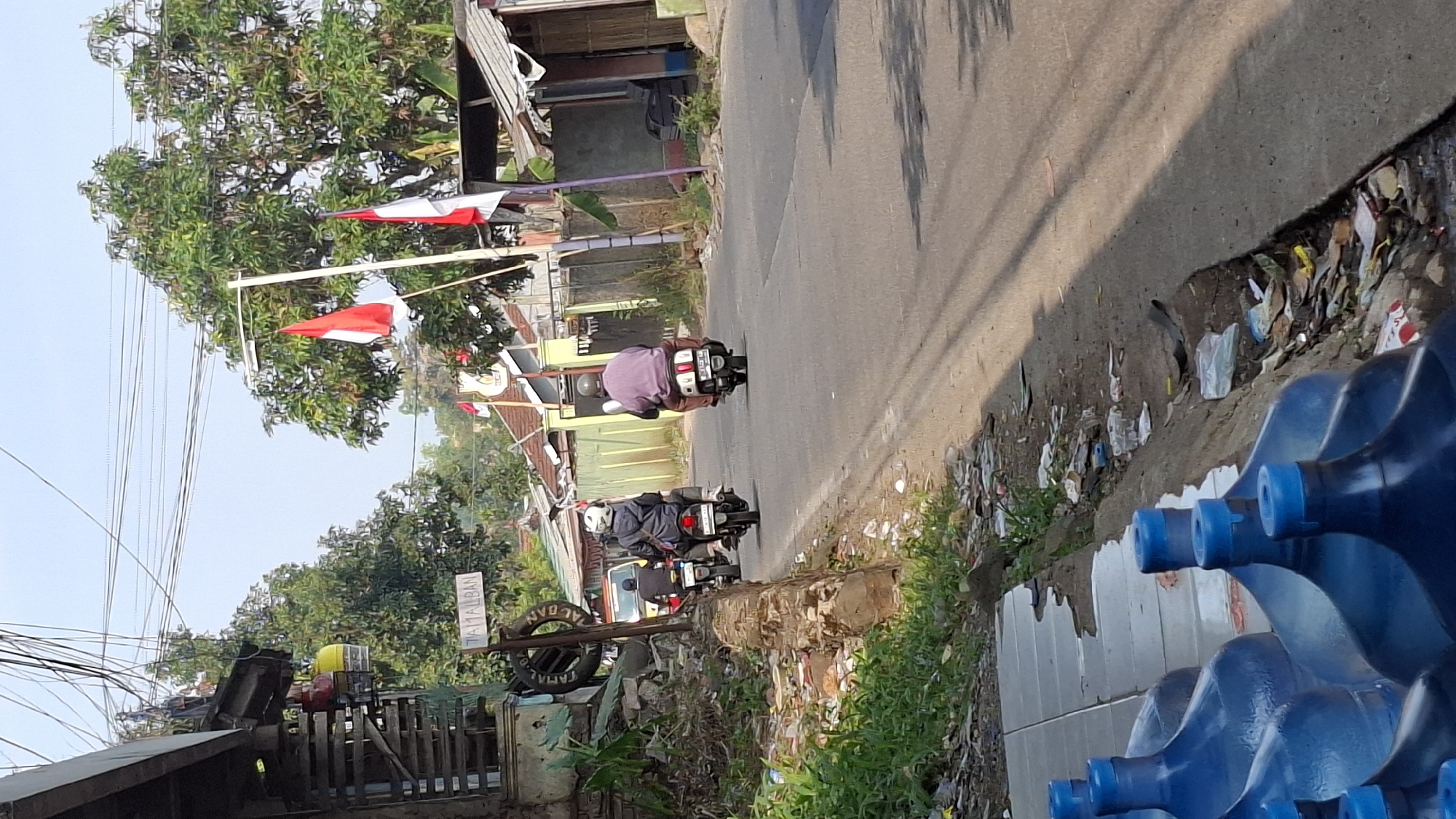 The image depicts a street scene in Cilaku, Indonesia. In the foreground, several large blue water bottles with blue caps are lined up on a tiled surface. To the right of the bottles is a dirt and gravel embankment littered with trash, including plastic bags, wrappers, and debris. This embankment slopes down to the edge of a paved road.
On the road, three motorcycles are visible, traveling away from the viewer. The first motorcycle, on the left, has two riders. The rider in front wears a white helmet and a dark jacket, while the passenger behind is not clearly visible. The second motorcycle, in the middle, also has two riders. The front rider wears a white helmet and a gray jacket. The passenger behind is not clearly visible. The third motorcycle, on the right, has a single rider wearing a light purple shirt and a dark helmet.
The environment includes buildings with corrugated metal roofs and fences along the roadside. Several Indonesian flags, red and white, are mounted on poles, indicating a festive or commemorative occasion. Lush green trees with dense foliage are present throughout the scene, suggesting a tropical climate. Overhead, a complex network of electrical wires crisscrosses the sky. A signpost with the text "TAMALBAN" is visible on the left side of the road, near a structure with what appear to be red and yellow gas canisters hanging from it. The overall impression is a somewhat cluttered but lived-in urban or semi-urban environment.